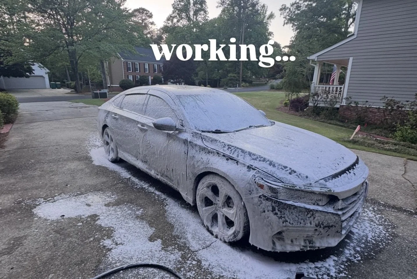 A grey car covered in soap suds during a car wash in a driveway, with the words 'working...' overlayed at the top.