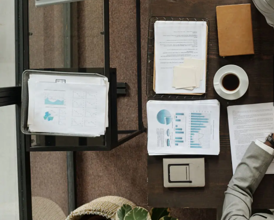 Top-down view of a cluttered office desk with various papers, charts, a coffee cup, a notebook, an electrical switch, and part of a person's arm.
