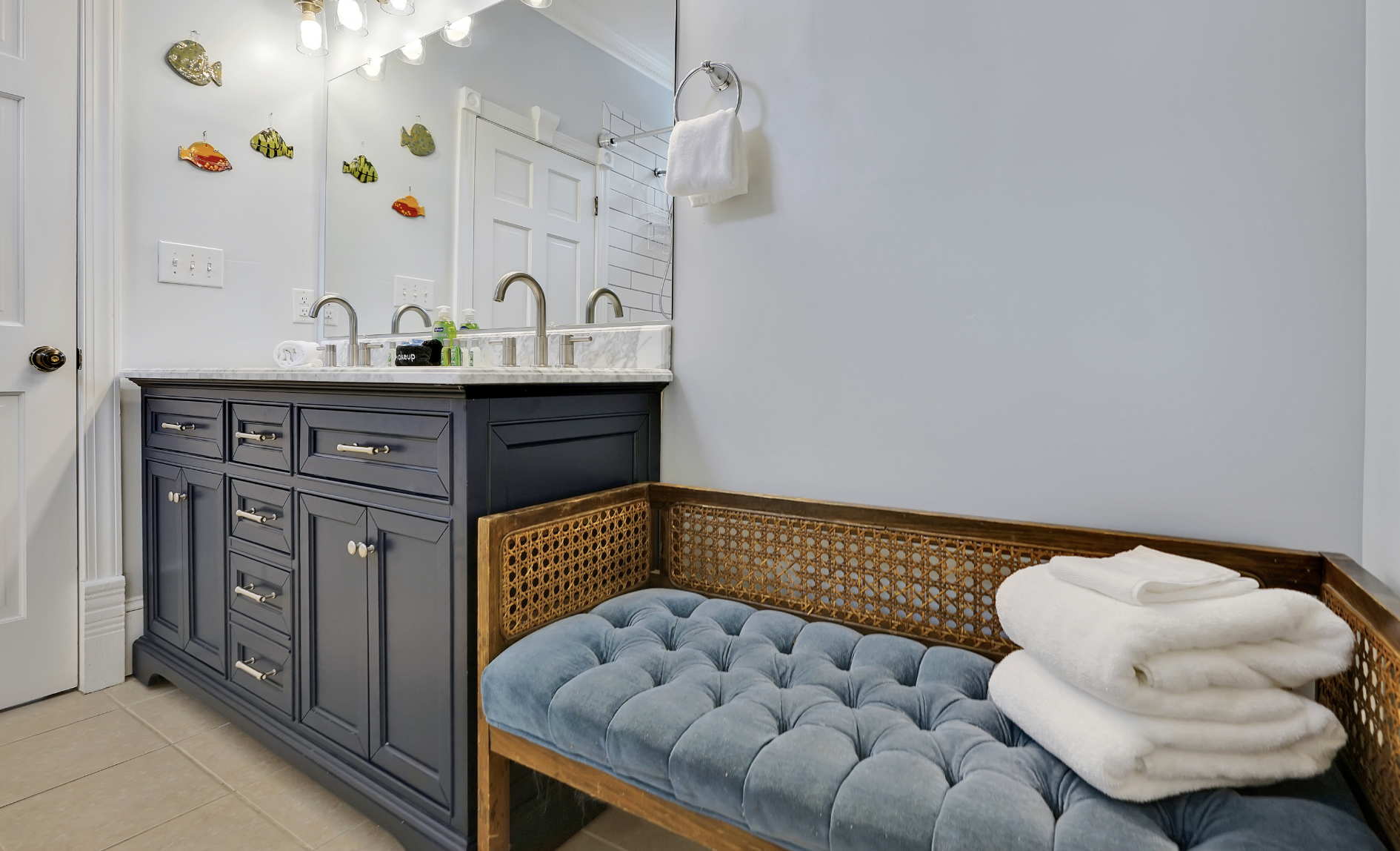 Spa-like bathroom in a rentable Wilmington, NC event home, featuring a navy blue double vanity with marble countertop and brushed nickel hardware, a tufted blue velvet and cane bench with fresh folded towels, globe vanity lighting, and whimsical fish