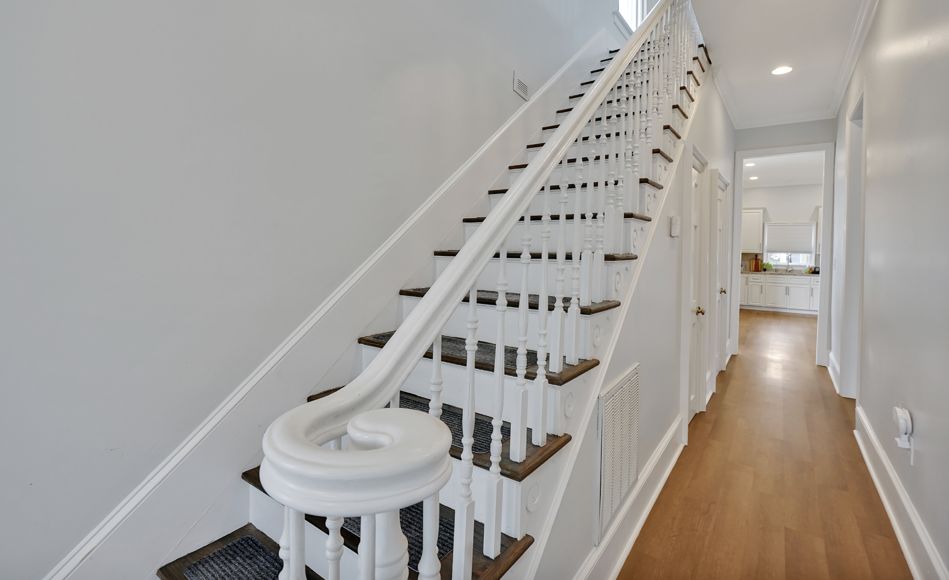 Elegant staircase and hallway in a rentable Wilmington, NC event home, featuring white spindle railings, dark wood treads with patterned carpet runners, warm hardwood floors, and a bright white hallway leading to the kitchen — perfect for wedding wee