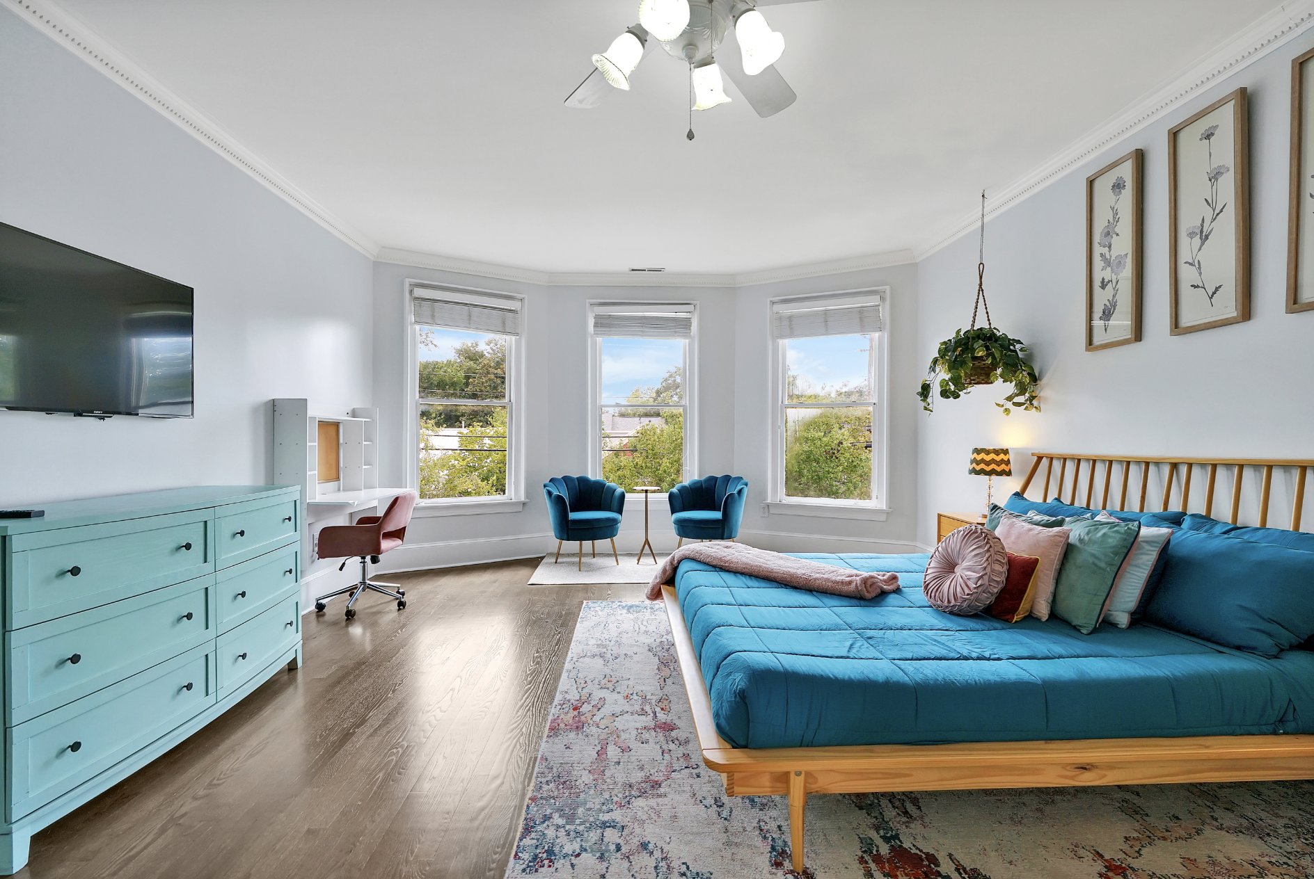 Spacious primary bedroom in a rentable Wilmington, NC event home, featuring a teal quilted bed with a natural wood frame, a teal dresser, two teal velvet accent chairs in a bay window seating area, a ceiling fan, hardwood floors, and a patterned area
