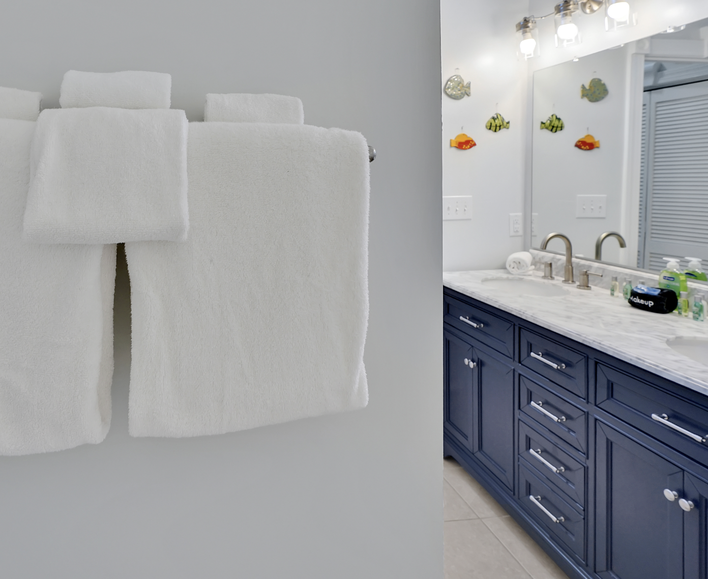 Well-appointed bathroom in a rentable Wilmington, NC event home, featuring fresh white towels on a towel bar and a double vanity with navy blue cabinetry, marble countertop, brushed nickel hardware, a large lighted mirror, and playful fish wall décor