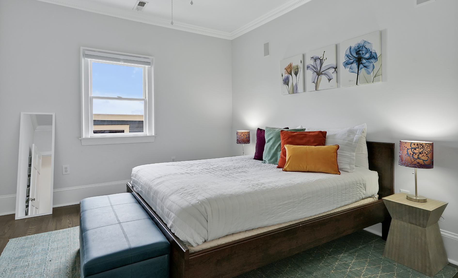 Cheerful guest bedroom in a rentable Wilmington, NC event home, featuring a dark wood platform bed with white bedding and colorful velvet throw pillows in orange, red, and teal, a teal upholstered bench, floral wall art, a patterned teal area rug, an