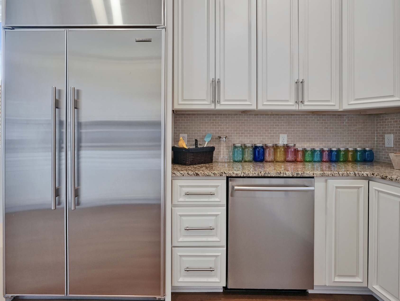 Kitchen detail in a rentable Wilmington, NC event home, showcasing a large stainless steel side-by-side refrigerator, stainless dishwasher, white shaker cabinets, granite countertops, gray subway tile backsplash, and a row of colorful mason jars — pe