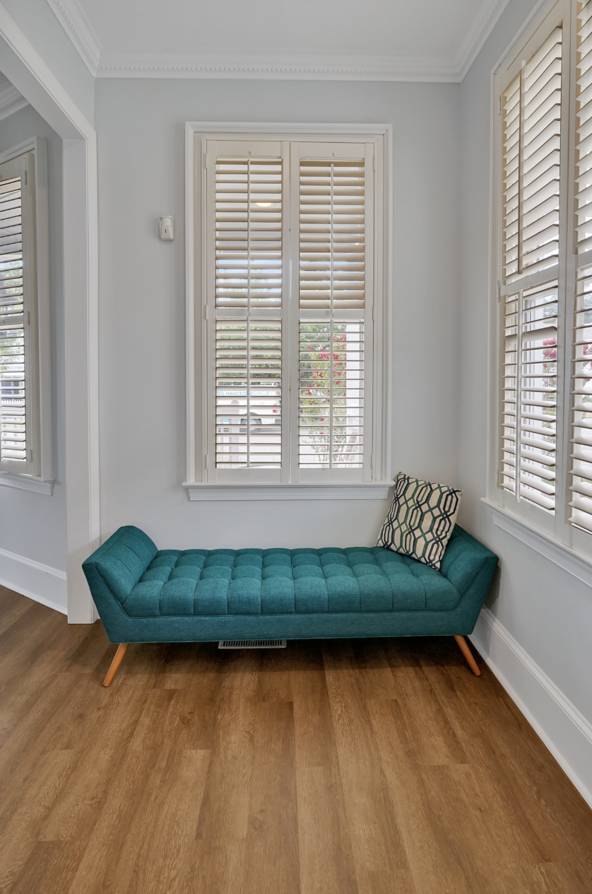 Cozy sitting nook in a rentable Wilmington, NC event home, featuring a tufted teal velvet chaise lounge with a geometric accent pillow, plantation shutters, light gray walls, and warm hardwood floors — perfect for wedding weekends, bridal showers, ba
