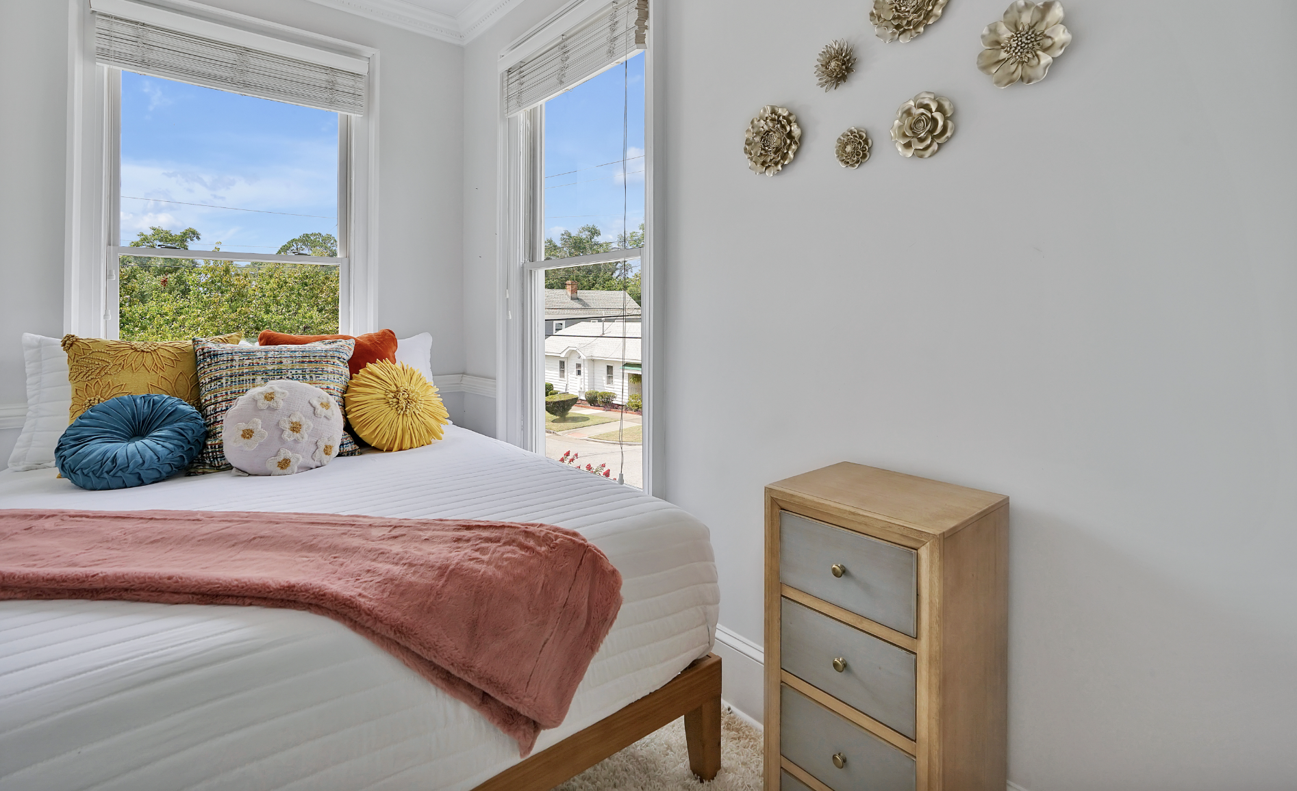 Bright and cheerful guest bedroom in a rentable Wilmington, NC event home, featuring a twin bed with white bedding, a pink throw, and colorful accent pillows in teal, yellow, and gold, a natural wood nightstand, gold floral wall décor, and a large wi