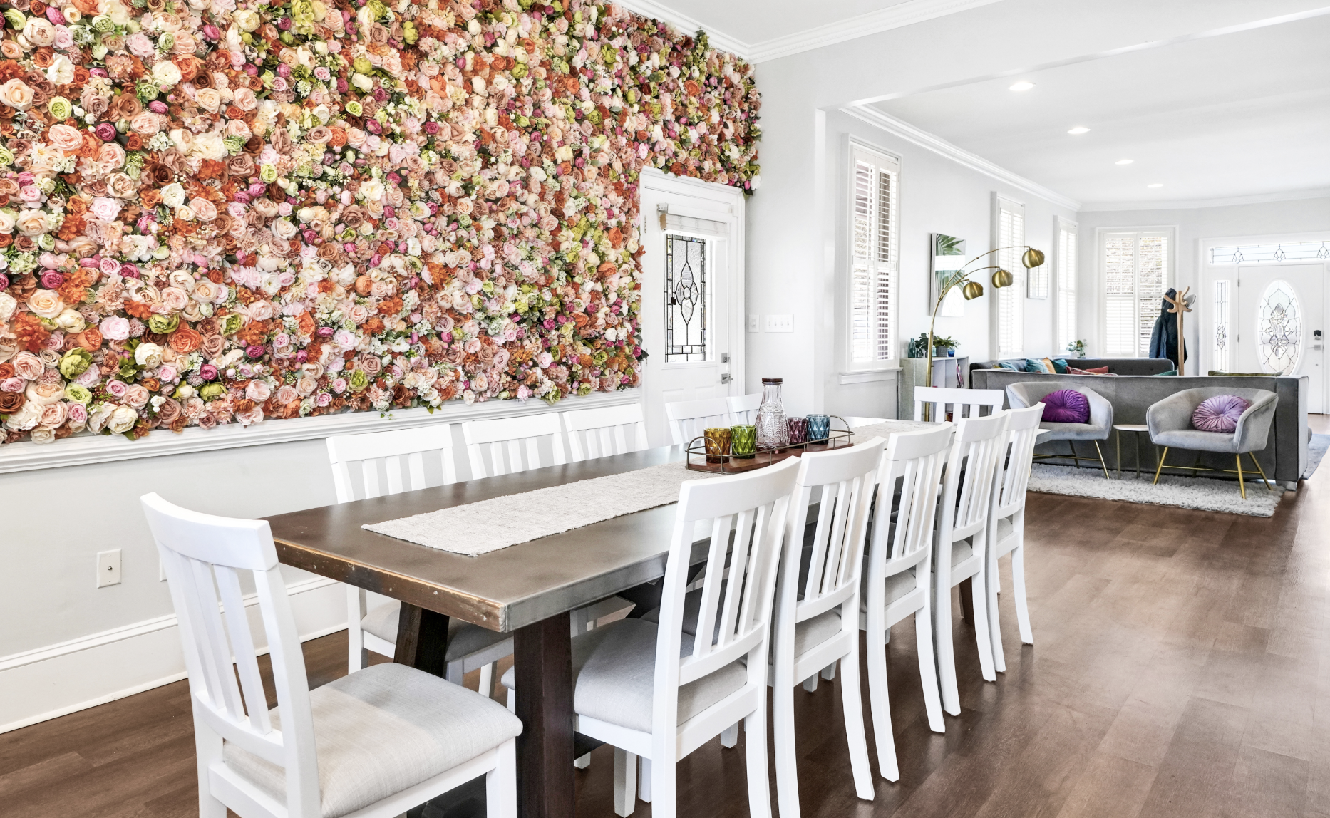 Stunning dining room in a rentable Wilmington, NC event home, featuring a long dark wood farmhouse table with white dining chairs, a dramatic floor-to-ceiling floral accent wall in blush, coral, and pink tones, dark hardwood floors, and an open view 