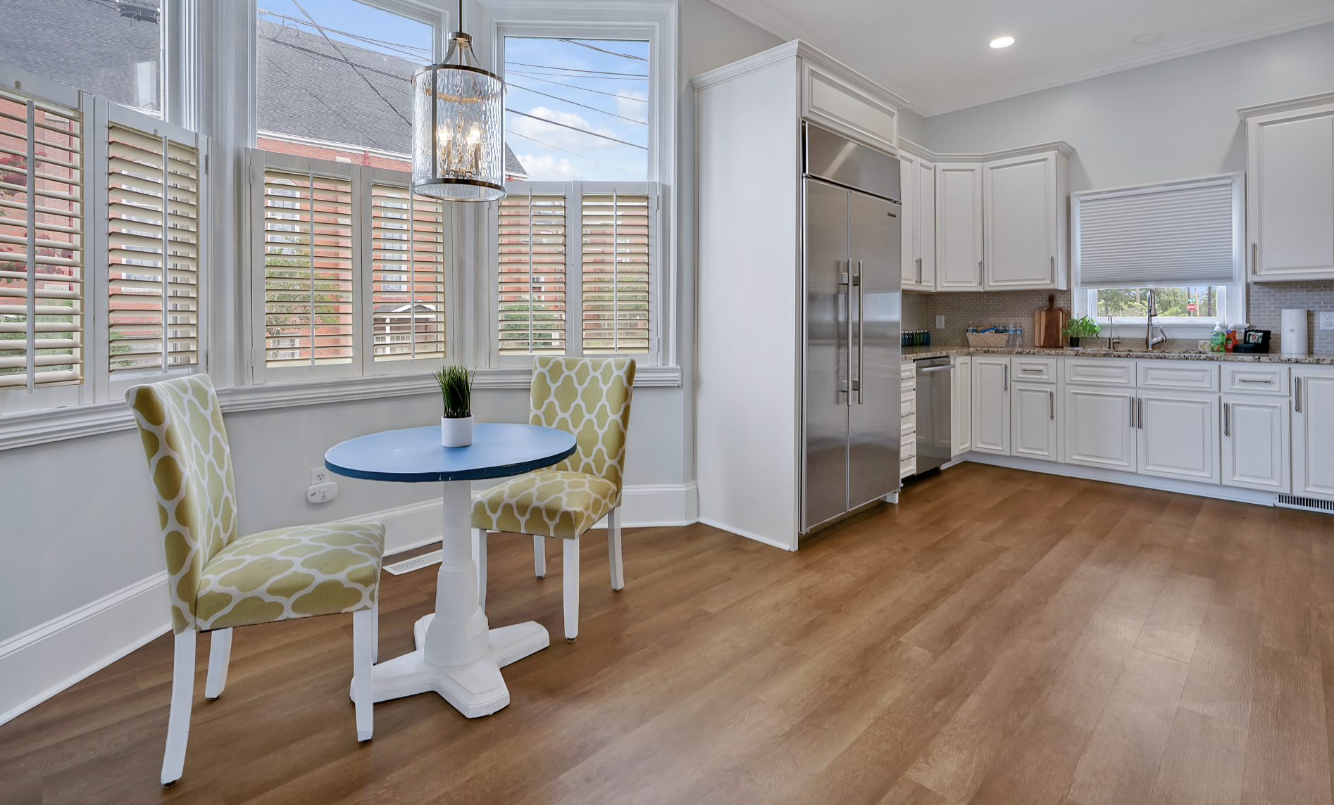 Sunny breakfast nook and kitchen in a rentable Wilmington, NC event home, featuring a small round blue café table with two green patterned upholstered chairs, a chandelier, plantation shutters, warm hardwood floors, and white cabinetry with stainless