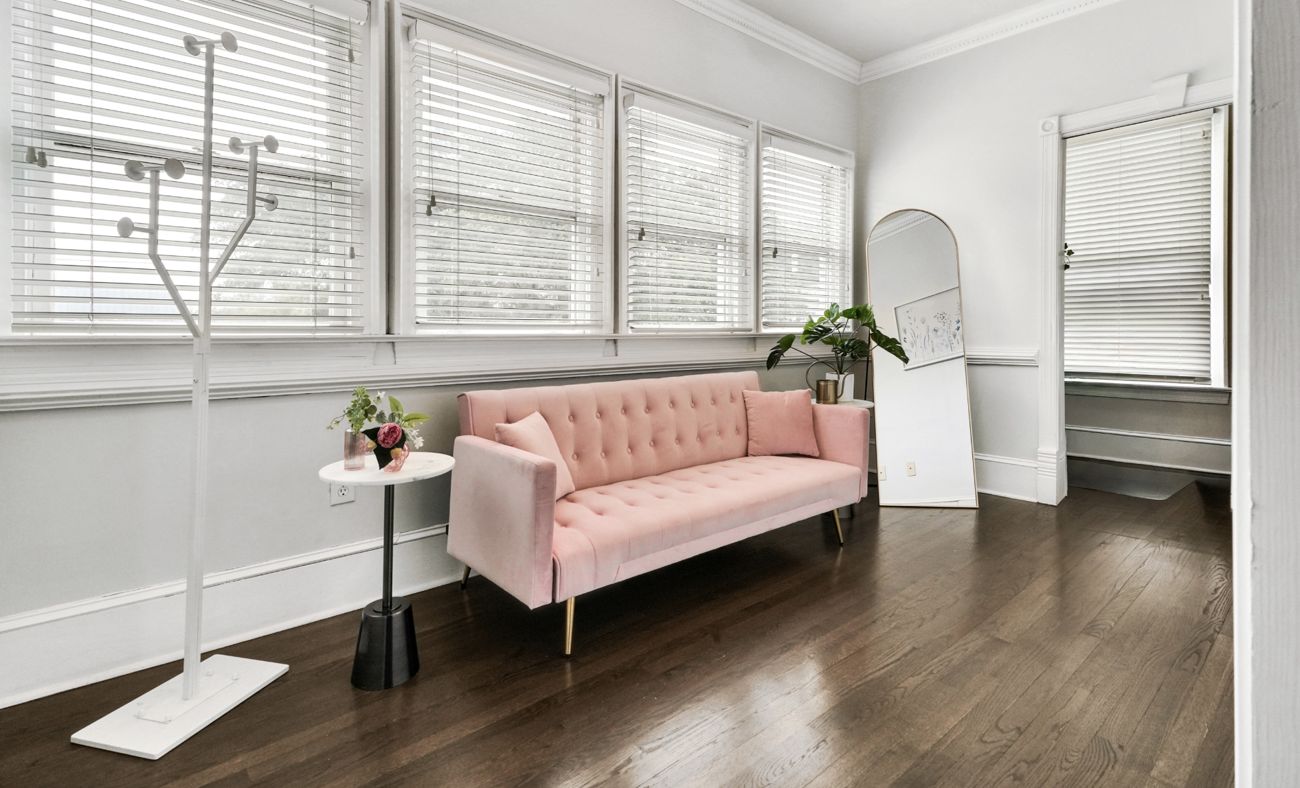 Chic dressing room or lounge area in a rentable Wilmington, NC event home, featuring a blush pink tufted velvet sofa with gold legs, a marble side table, a tall arched floor mirror, a decorative coat rack, dark hardwood floors, and white plantation s