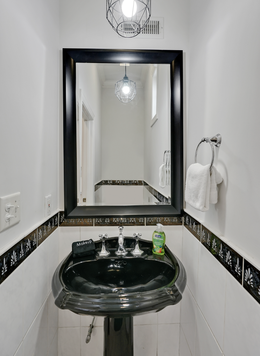 Stylish powder room in a rentable Wilmington, NC event home, featuring a dramatic black pedestal sink, a large black-framed mirror, a decorative tile border, white walls, and an industrial-style pendant light — perfect for wedding weekends, bridal sh