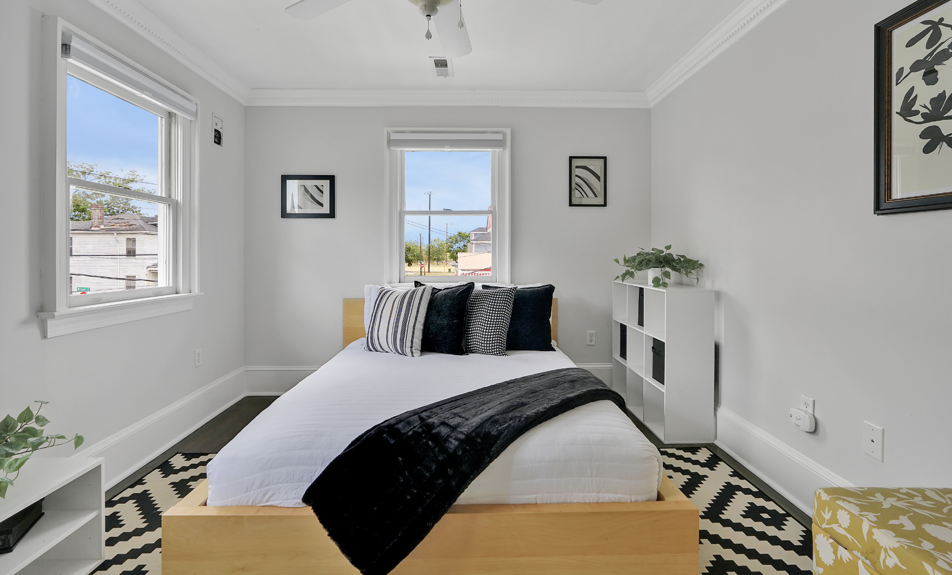 Modern black-and-white guest bedroom in a rentable Wilmington, NC event home, featuring a low wood platform bed with white bedding and black accent pillows, a bold geometric black-and-white area rug, light gray walls, crown molding, and natural light