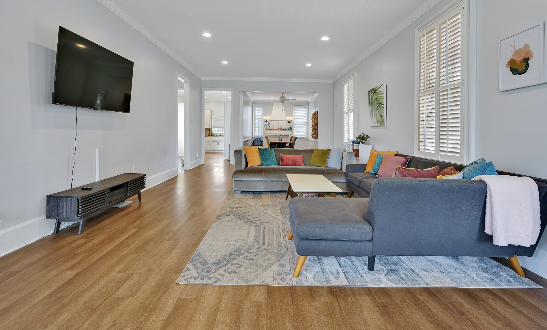 Bright and airy living room in a rentable Wilmington, NC event home, featuring a charcoal gray sectional sofa with colorful throw pillows in teal, yellow, orange, and olive, a beige curved sofa, white plantation shutters, light blue walls, and a patt