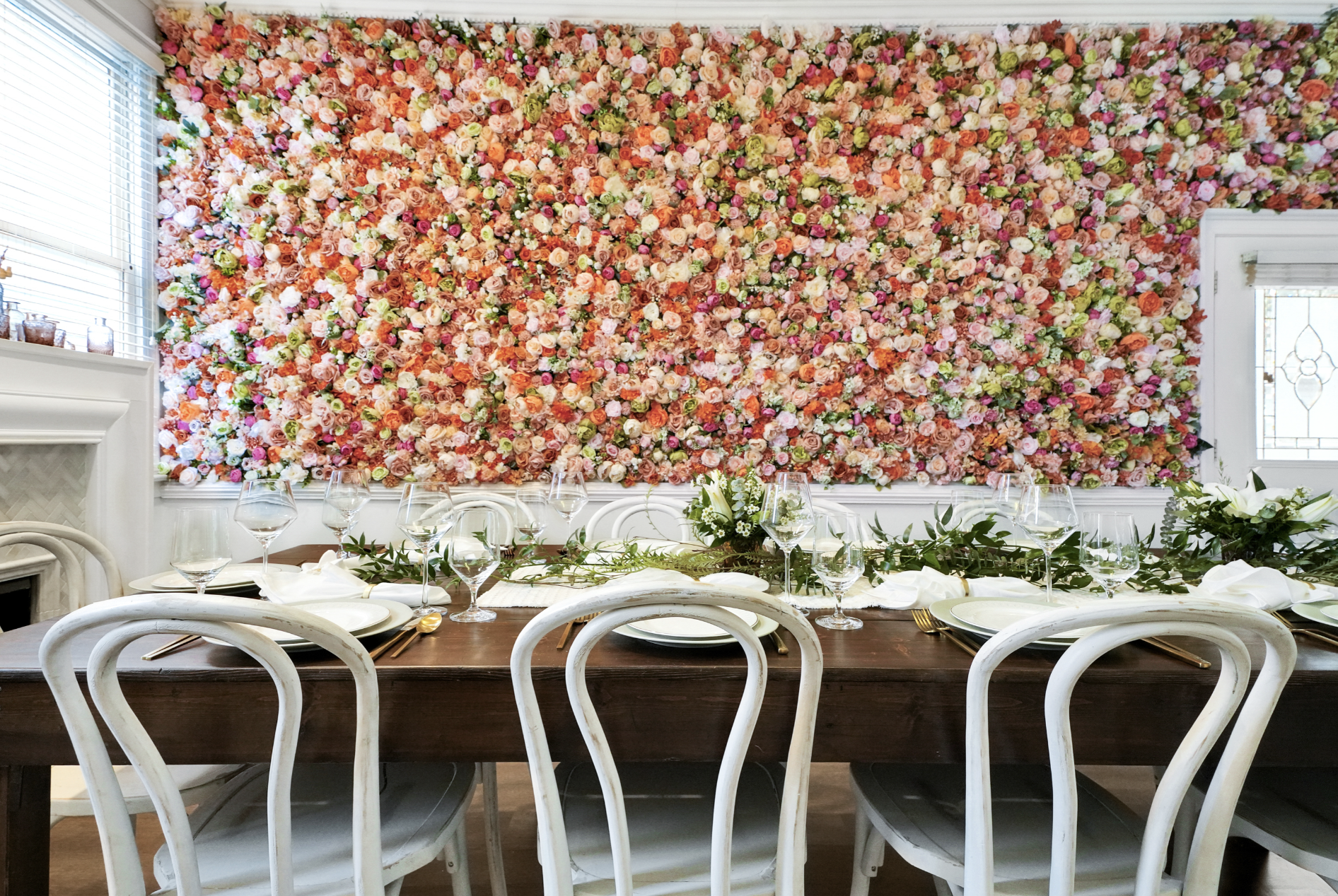 Elegantly set dining table in a rentable Wilmington, NC event home, with white cross-back chairs, white plates, crystal stemware, and a lush greenery runner centerpiece set against a dramatic floor-to-ceiling floral wall in blush and coral — perfect 