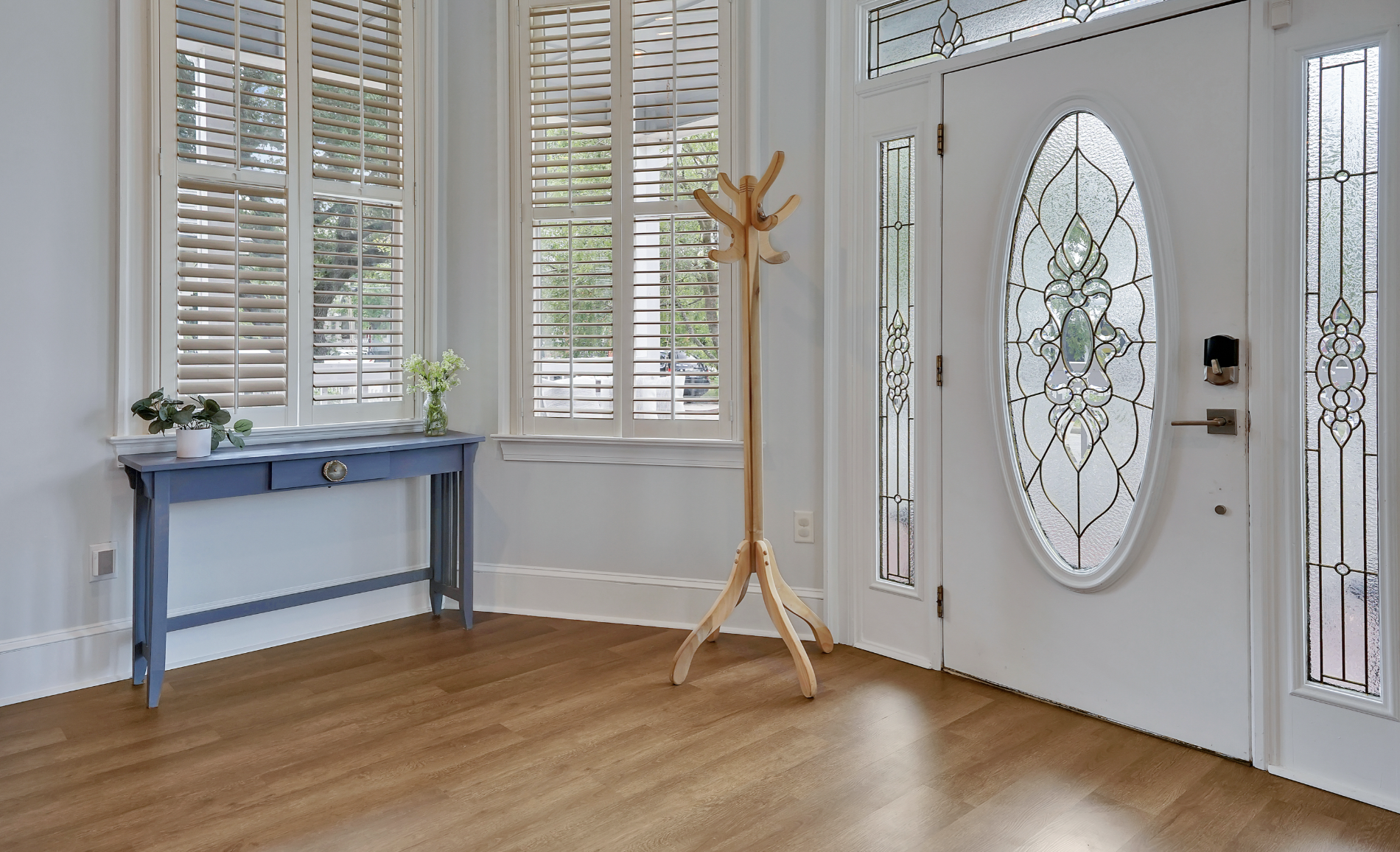 Welcoming entryway of a rentable Wilmington, NC event home, featuring a white front door with an ornate oval leaded glass insert, a natural wood coat rack, a blue console table with fresh florals, plantation shutters, and warm hardwood floors — perfe
