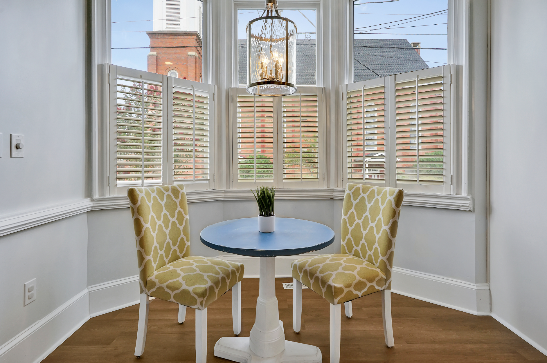 Charming bay window breakfast nook in a rentable Wilmington, NC event home, featuring a small round blue café table, two yellow patterned upholstered chairs, a beaded chandelier, plantation shutters, and warm hardwood floors — perfect for wedding wee