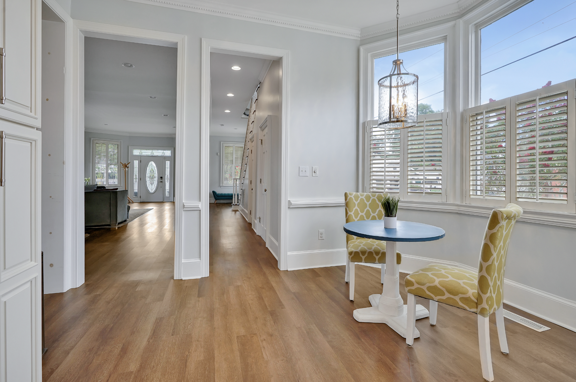 Open, light-filled interior of a rentable Wilmington, NC event home, showing a cozy bay window breakfast nook with a blue café table and green patterned chairs, a gold chandelier, plantation shutters, hardwood floors, and a view through the hallway t