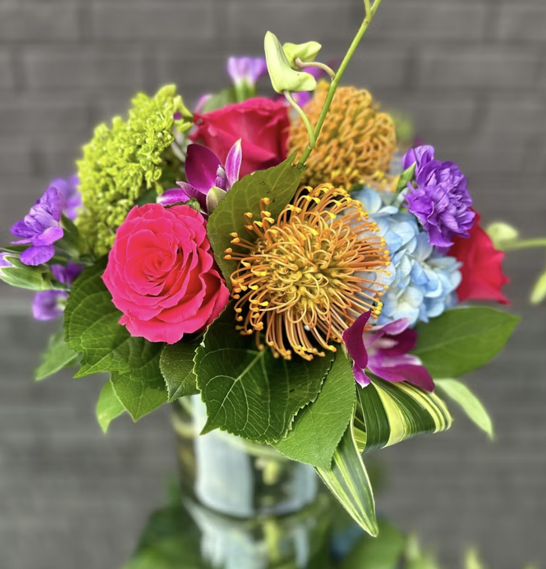 Image 1: Vibrant mixed floral arrangement featuring a hot pink rose, orange pincushion protea, blue hydrangea, purple orchids, and chartreuse greenery — available as an add-on for events at a rentable Wilmington, NC event home, perfect for wedding w