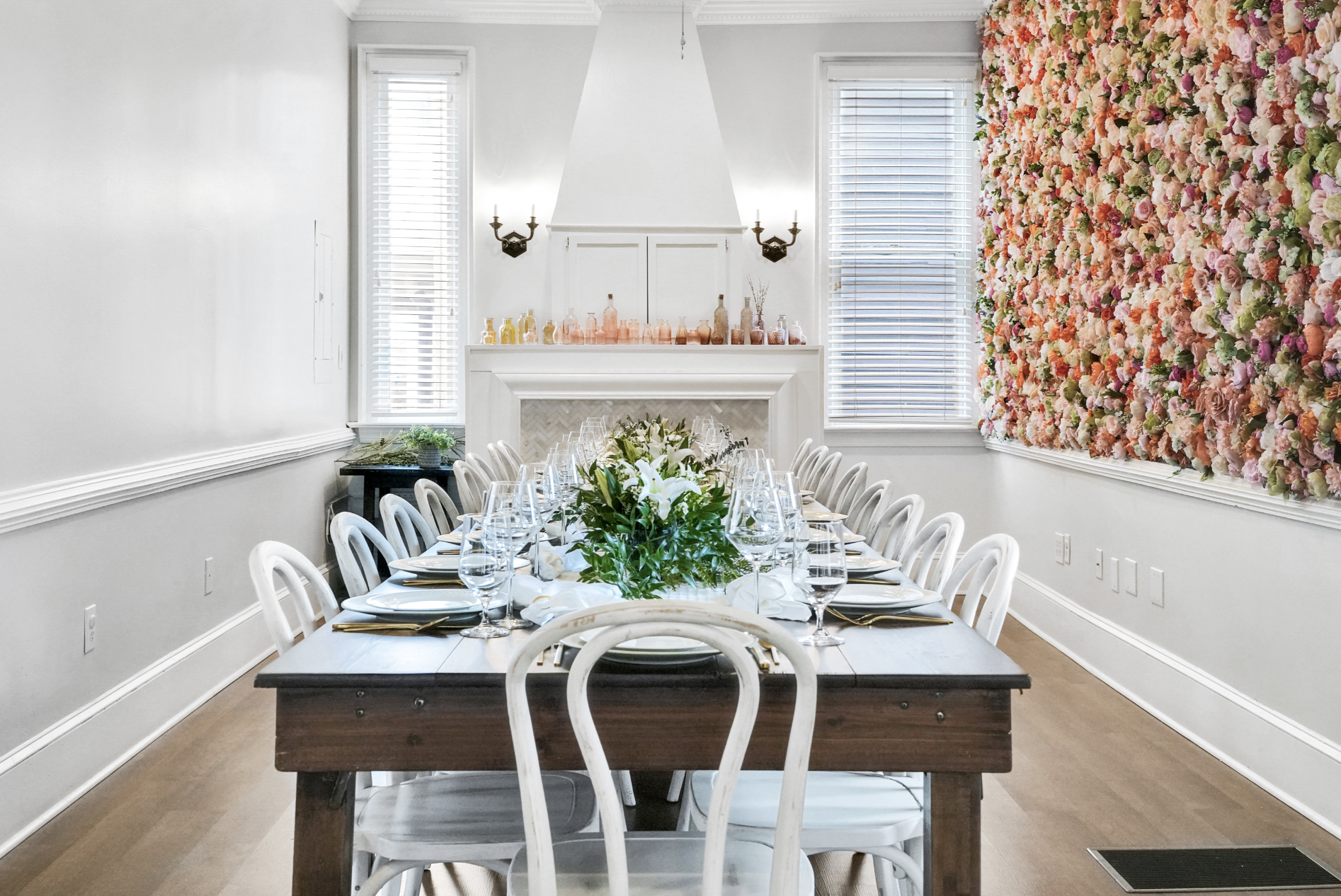 Fully set dining room in a rentable Wilmington, NC event home, featuring a long farmhouse table with white chairs, crystal glassware, a fresh floral centerpiece, a white fireplace with colorful bottle décor, and a floor-to-ceiling floral accent wall 