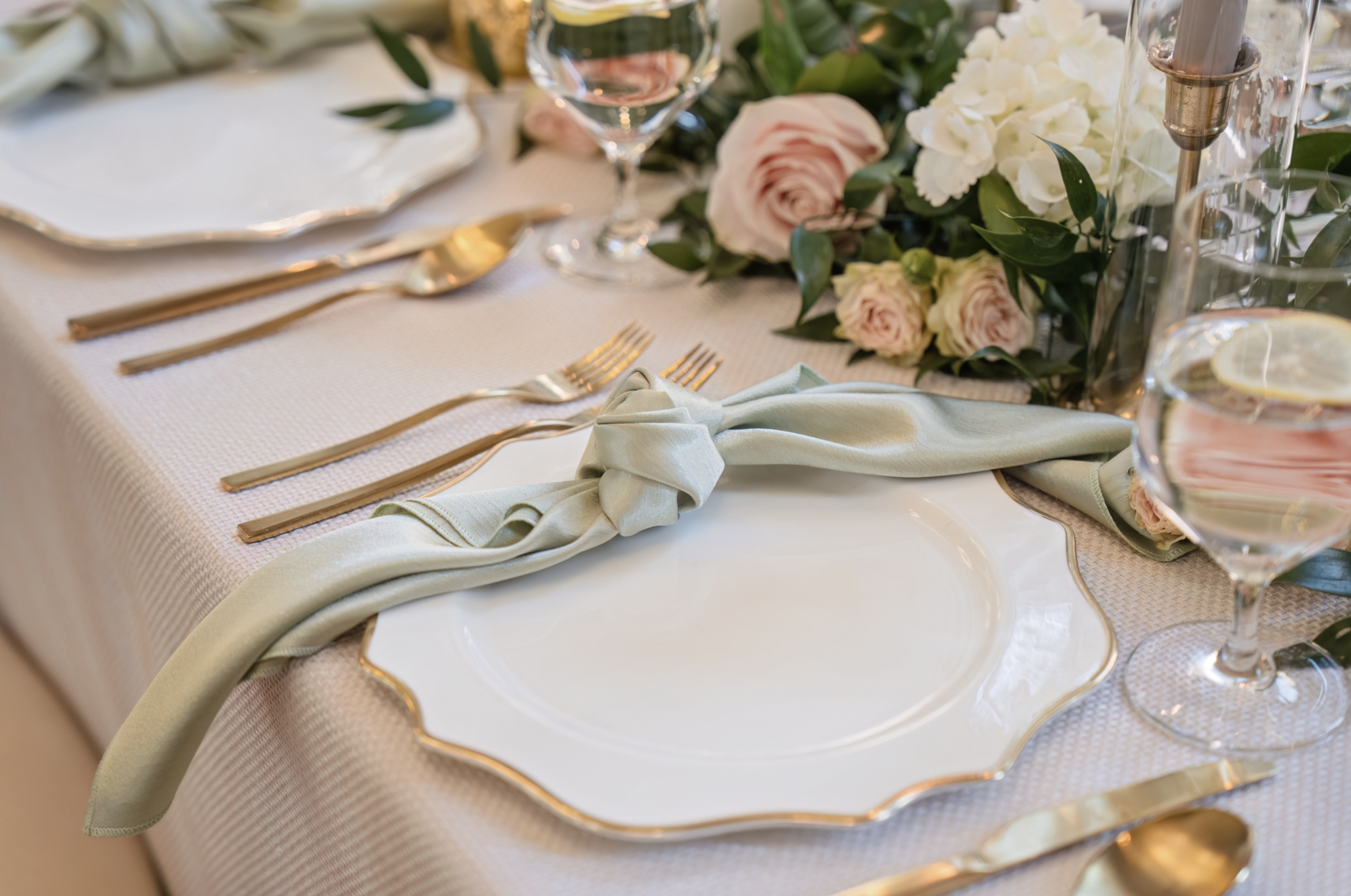 Elegant place setting featuring a white scalloped plate with gold rim, a sage green knotted linen napkin, gold flatware, crystal stemware, and a blush rose and white hydrangea floral centerpiece — available as a tablescape add-on for events at a rent