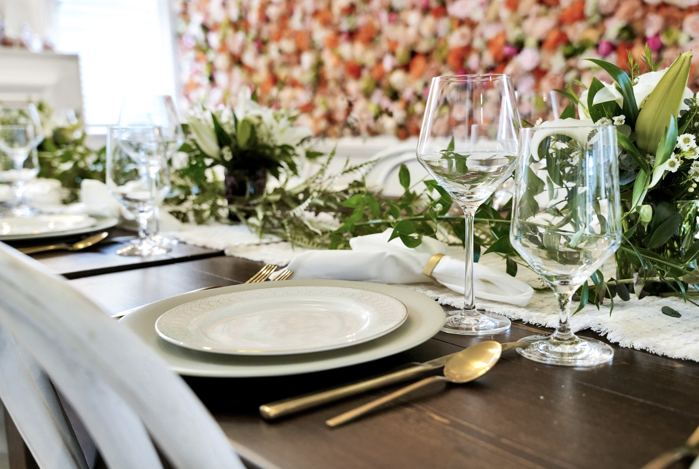 Elegant place setting detail in a rentable Wilmington, NC event home, featuring stacked white plates, gold flatware, crystal wine glasses, a linen napkin, and lush greenery centerpiece against the floral accent wall — perfect for wedding weekends, br