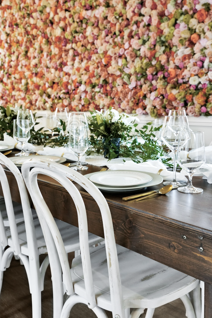 Side view of a beautifully set dining table in a rentable Wilmington, NC event home, with white cross-back chairs, gold flatware, crystal stemware, and a greenery runner centerpiece set against the signature floor-to-ceiling floral wall — perfect for