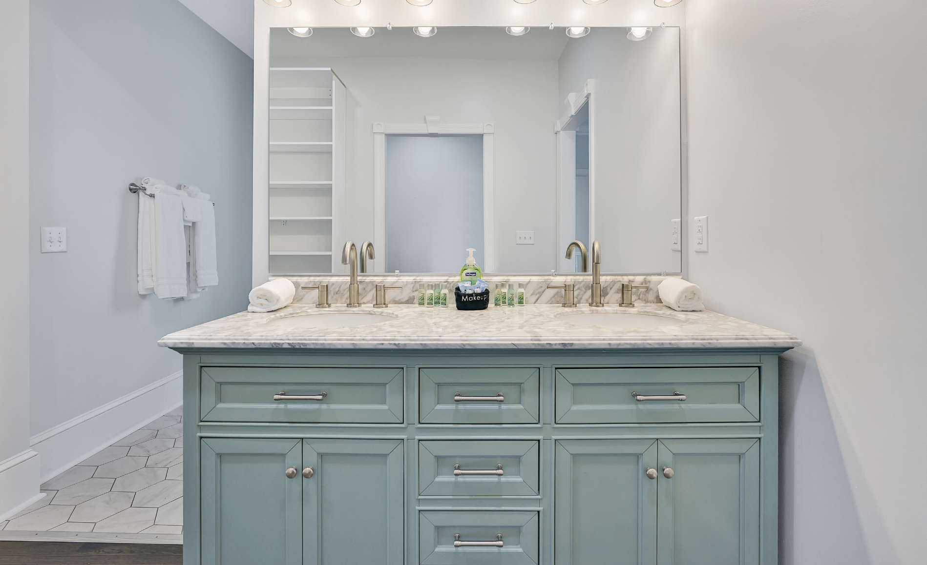 Elegant bathroom in a rentable Wilmington, NC event home, featuring a double vanity with a sage green cabinet, marble countertop, brushed nickel fixtures, a large mirror with Hollywood-style lighting, built-in shelving, and geometric tile flooring — 