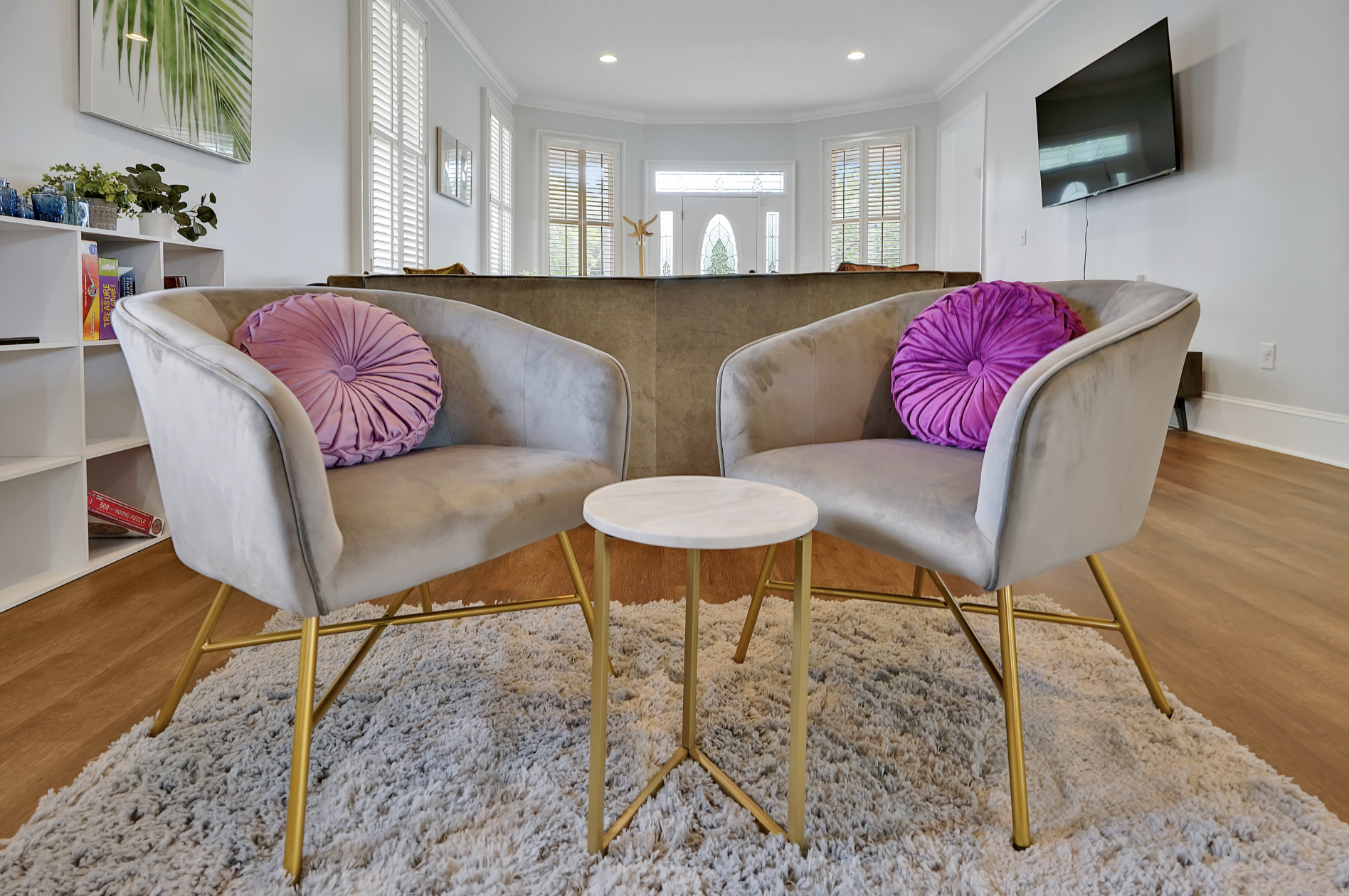 Stylish sitting area in a rentable Wilmington, NC event home, featuring two gray velvet bucket chairs with gold legs and pink pleated throw pillows, a small round marble side table, a plush cream shag rug, and plantation shutters — perfect for weddin
