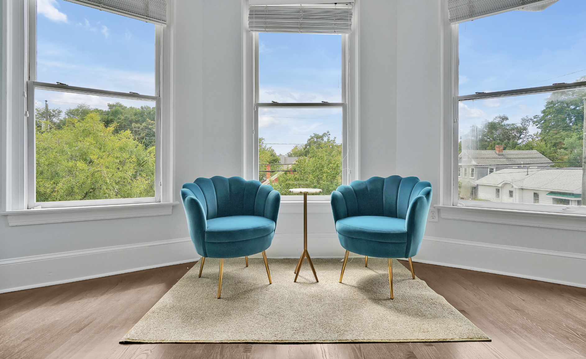 Bright bay window seating area in a rentable Wilmington, NC event home, featuring two teal velvet scalloped accent chairs with gold legs, a small gold side table, a cream shag rug, and sweeping tree-top views — perfect for wedding weekends, bridal sh