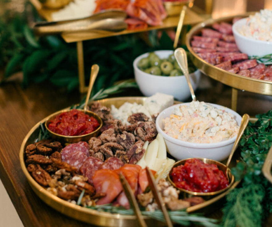 Abundant grazing board display with gold serving trays and bowls filled with cured meats, candied pecans, pimento cheese, tomato jam, olives, and assorted cheeses, surrounded by fresh greenery — available as a catering add-on for events at a rentable