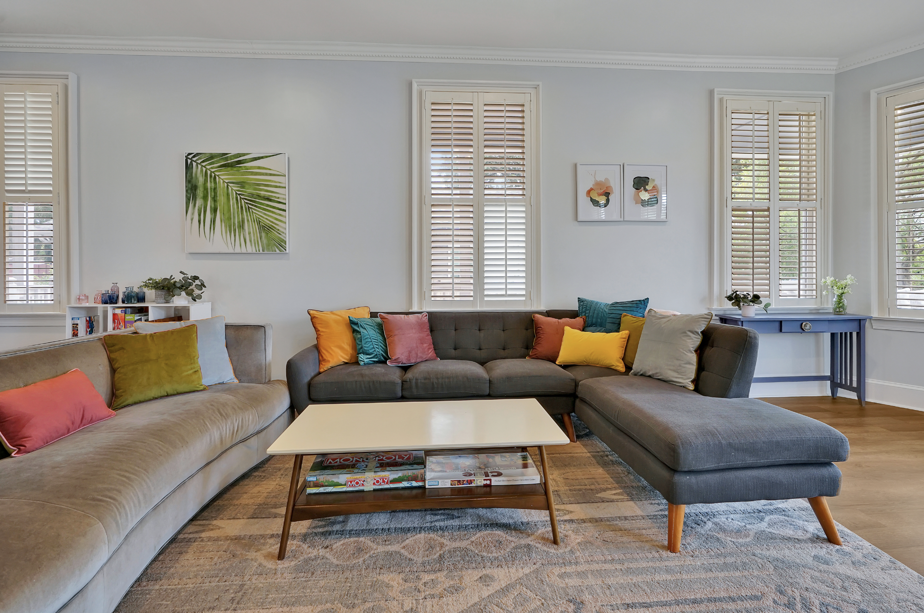 Bright and airy living room in a rentable Wilmington, NC event home, featuring a charcoal gray sectional sofa with colorful throw pillows in teal, yellow, orange, and olive, a beige curved sofa, white plantation shutters, light blue walls, and a patt