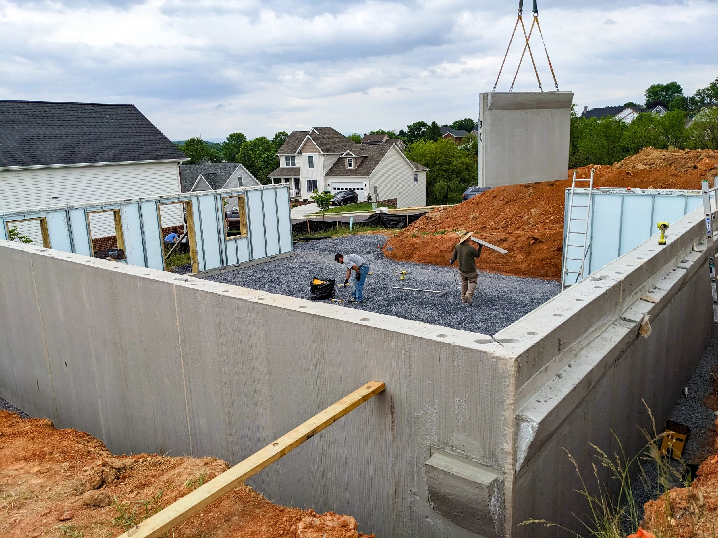 Construction site with a crane lifting a concrete wall panel, with residential houses and trees in the background.