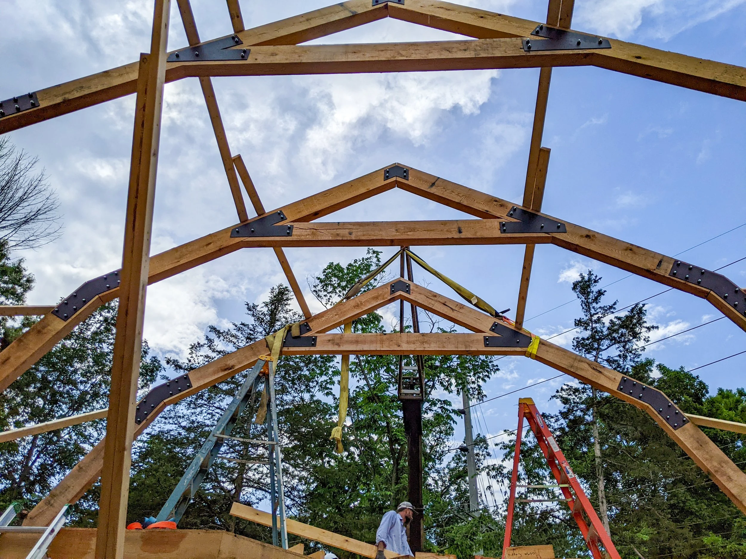 Wooden framework for a building under construction, with ladders, construction tools, and workers, set outdoors with trees and a partly cloudy sky.