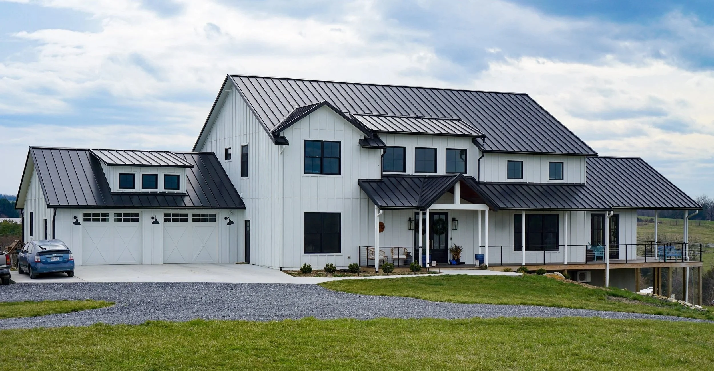 A modern white farmhouse with a metal roof, garage, and porch, surrounded by green grass and a partly cloudy sky.