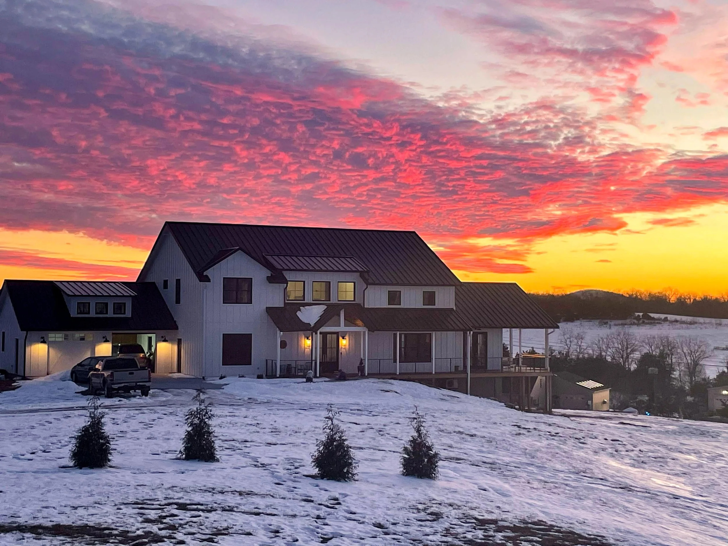 A residential house with a dark roof and lights glowing inside and outside, set against a vibrant sunset sky, with snow on the ground.