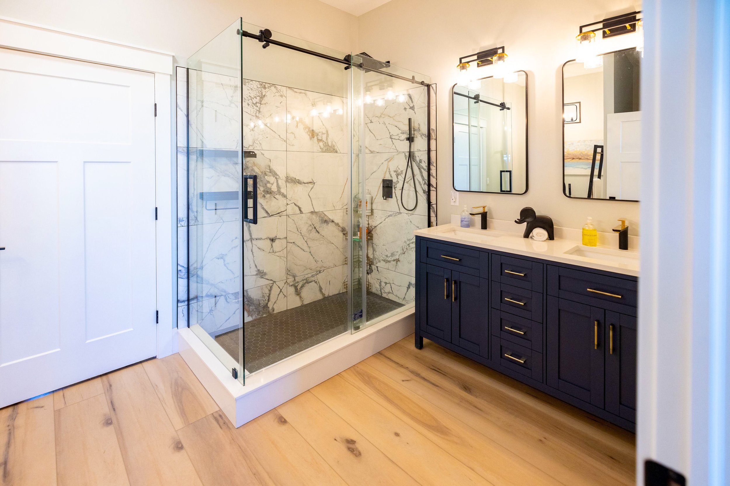 Bathroom with a modern glass-enclosed shower, white walls, and a blue vanity. Two mirrors and lighting fixtures above the sink, with wood flooring.