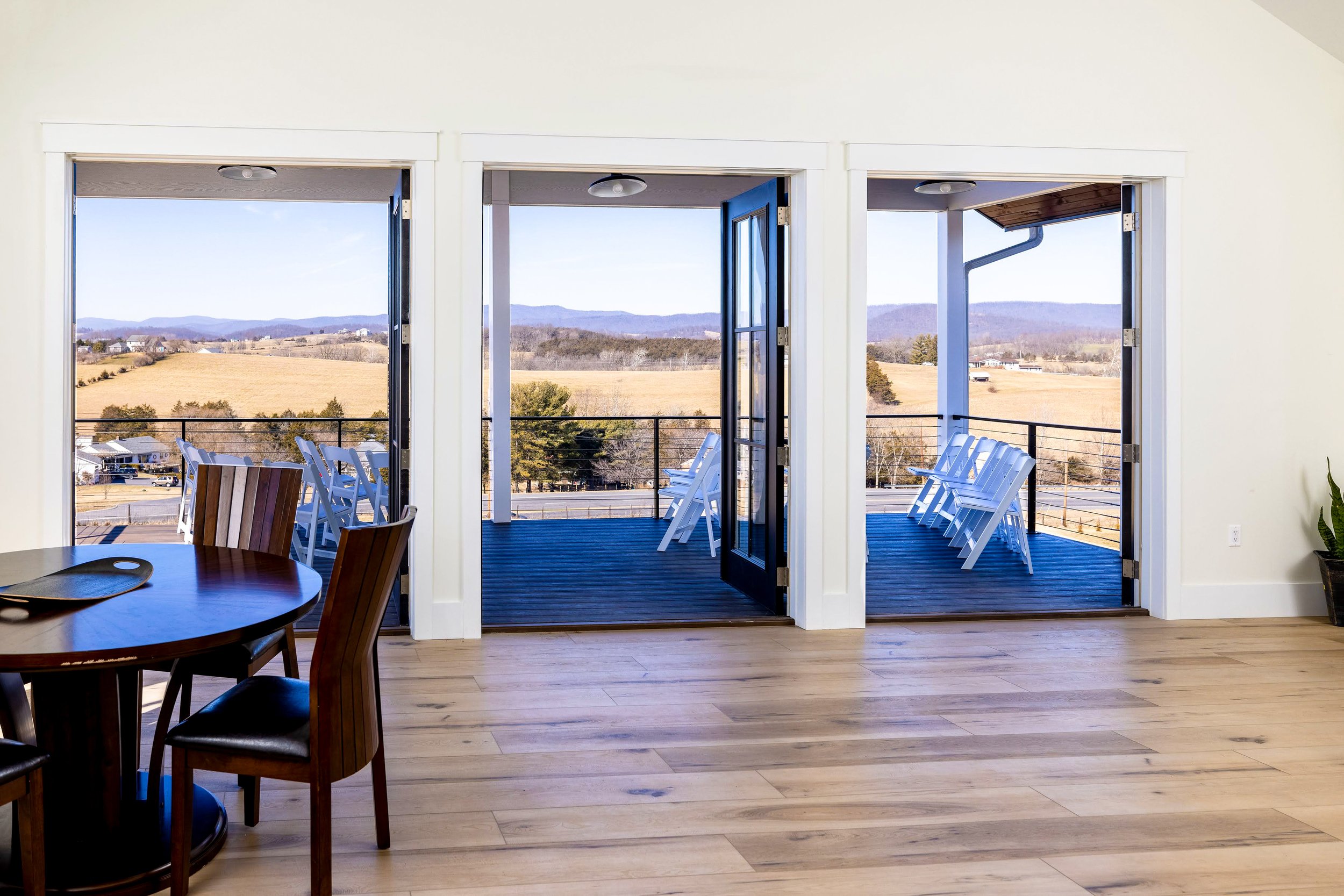 Interior view of a room with a sliding glass door opening to a balcony. The balcony has white chairs, overlooking a rural landscape with fields and mountains in the distance.