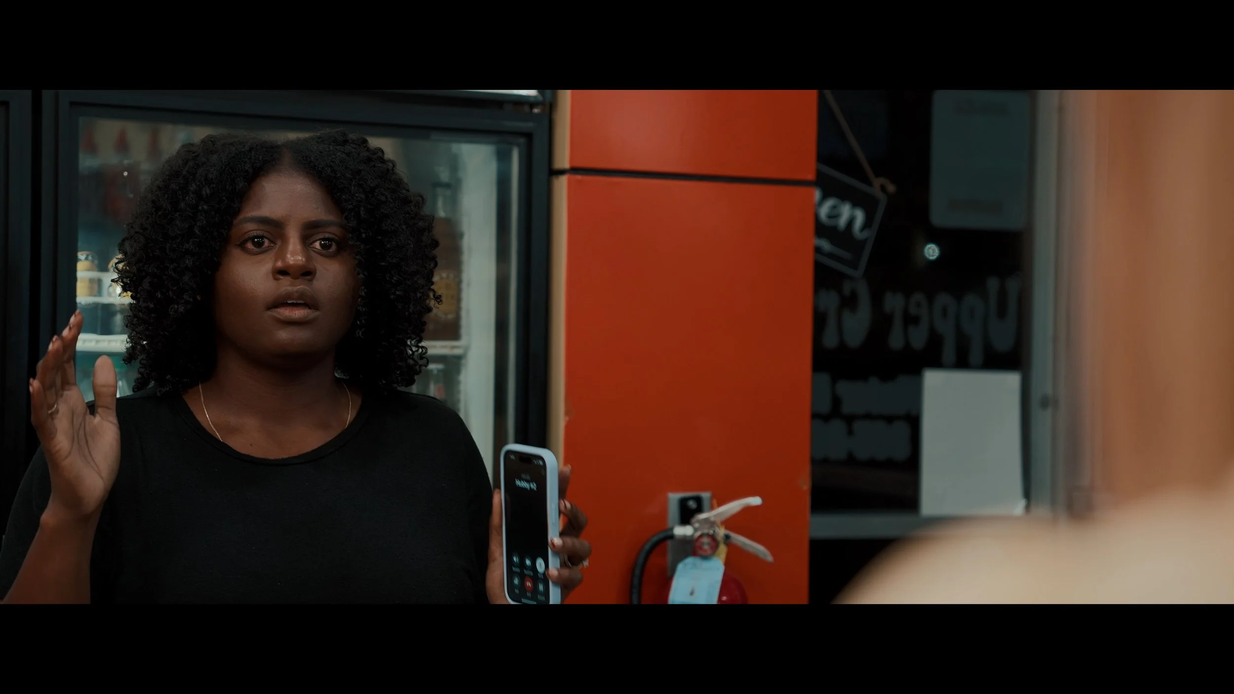 A woman with dark, curly hair is standing in front of a refrigerator with a glass door, inside which drinks are visible. She is raising her right hand and holding a phone in her left hand.