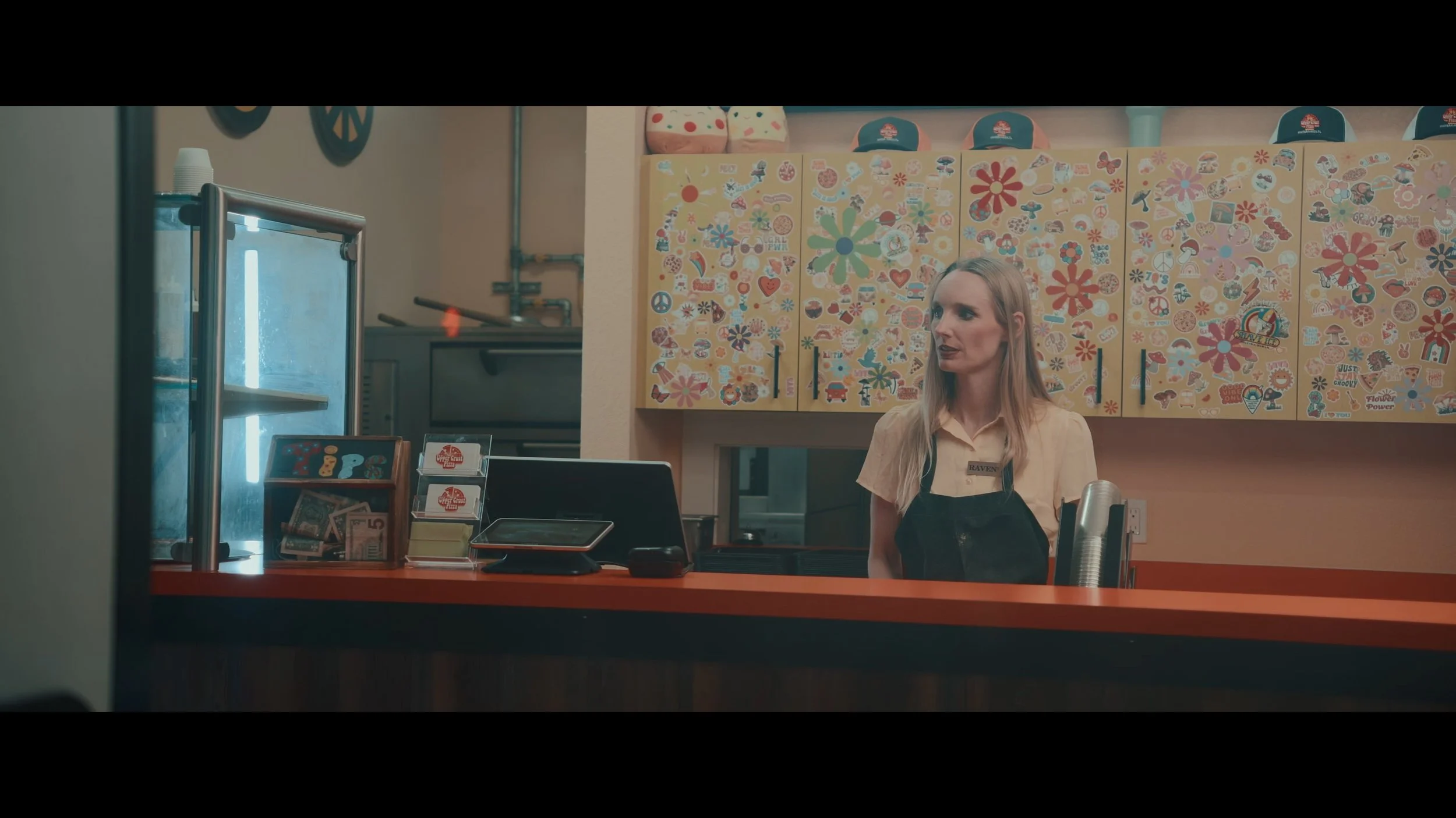 A woman with long blonde hair wearing a beige shirt and black apron standing behind a counter in a colorful ice cream shop decorated with stickers of flowers, peace signs, rainbows, and various playful images.