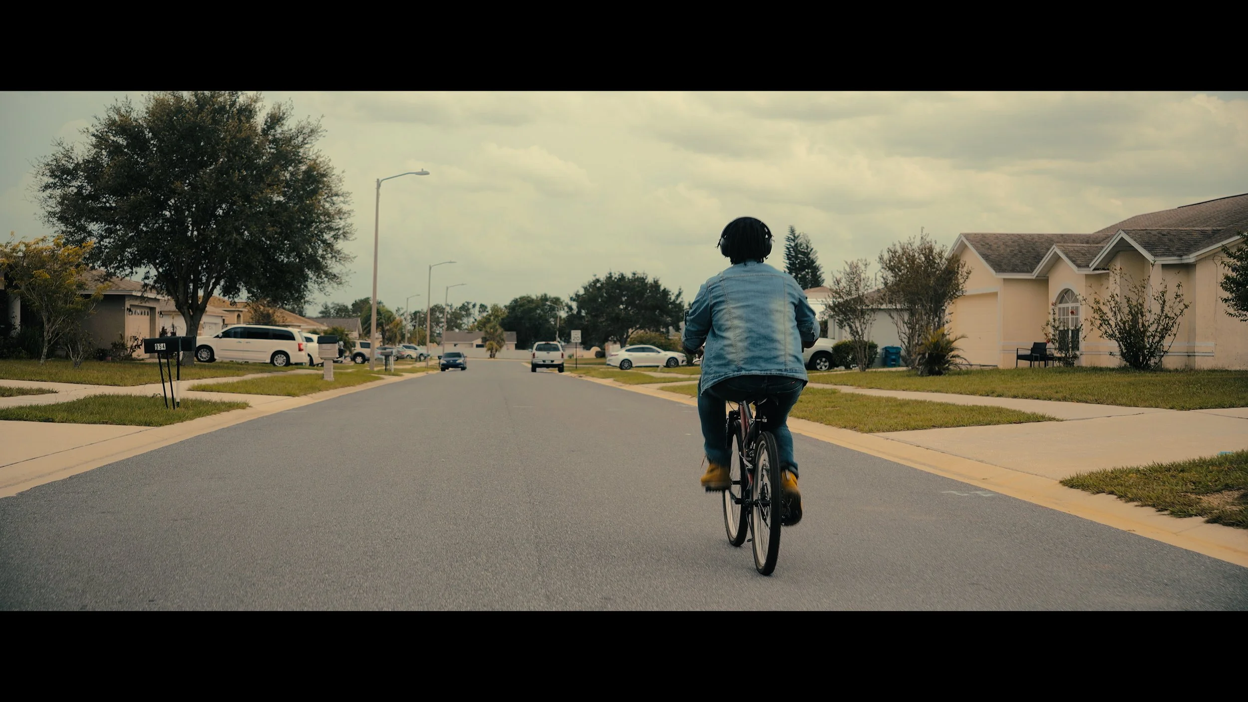 A person riding a bicycle down a suburban street with houses and cars on a cloudy day.