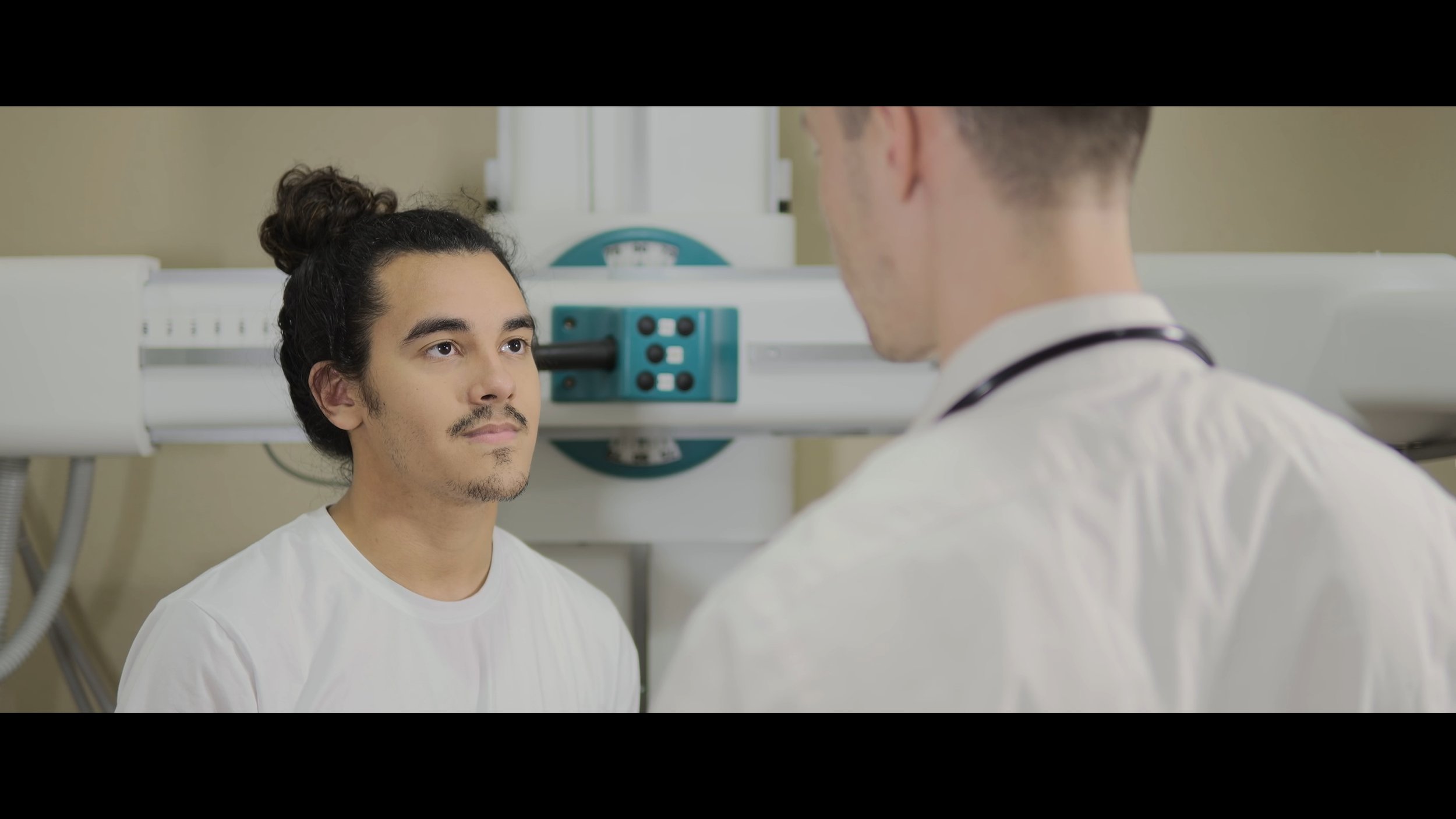 A male patient sitting in a medical examination room looking at a doctor, who is standing in front of him and wearing a white coat and stethoscope.