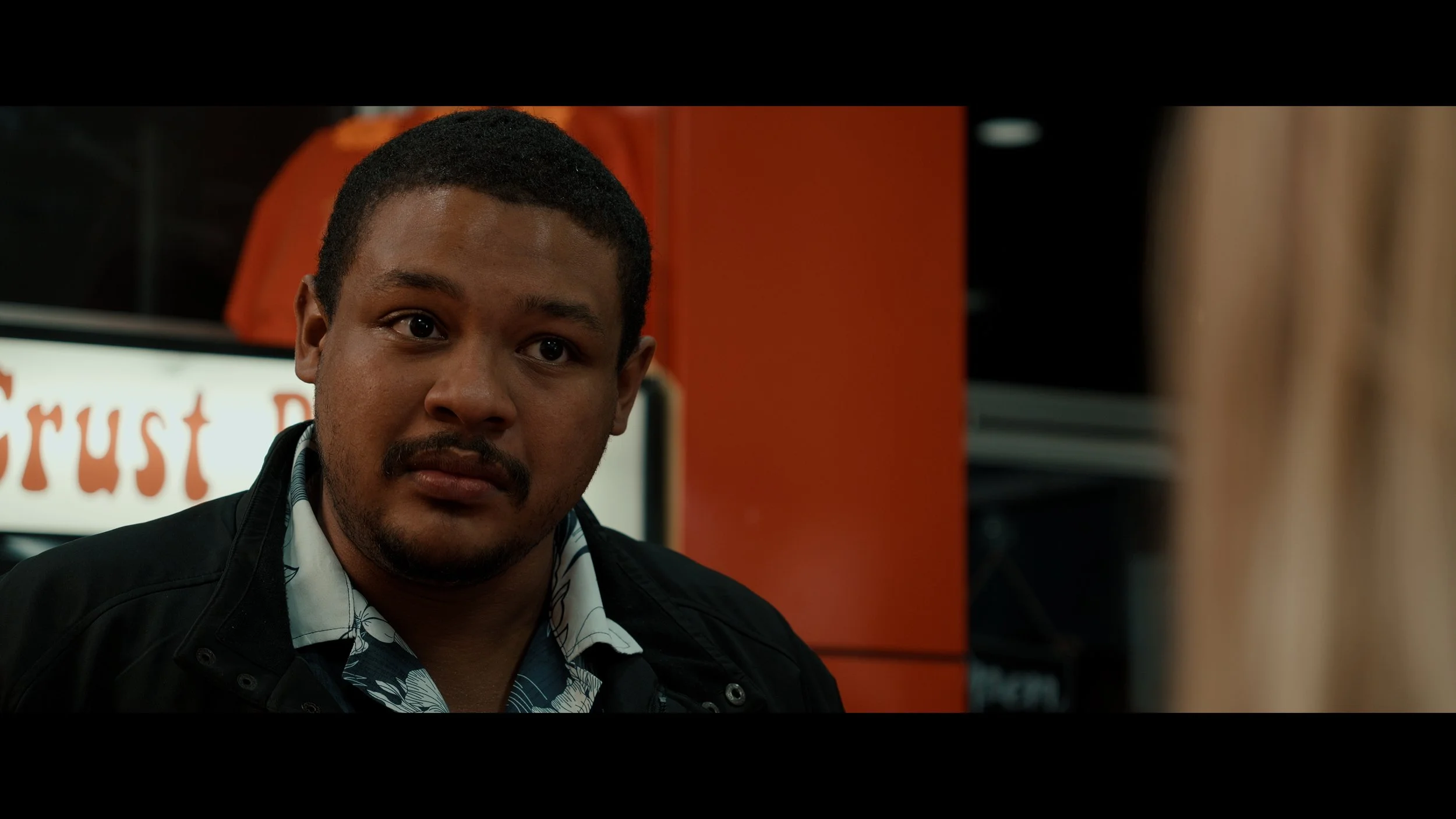 A man with short black hair, a mustache, and a goatee wearing a patterned shirt and a black jacket, standing in front of an orange wall with a partially visible sign behind him.