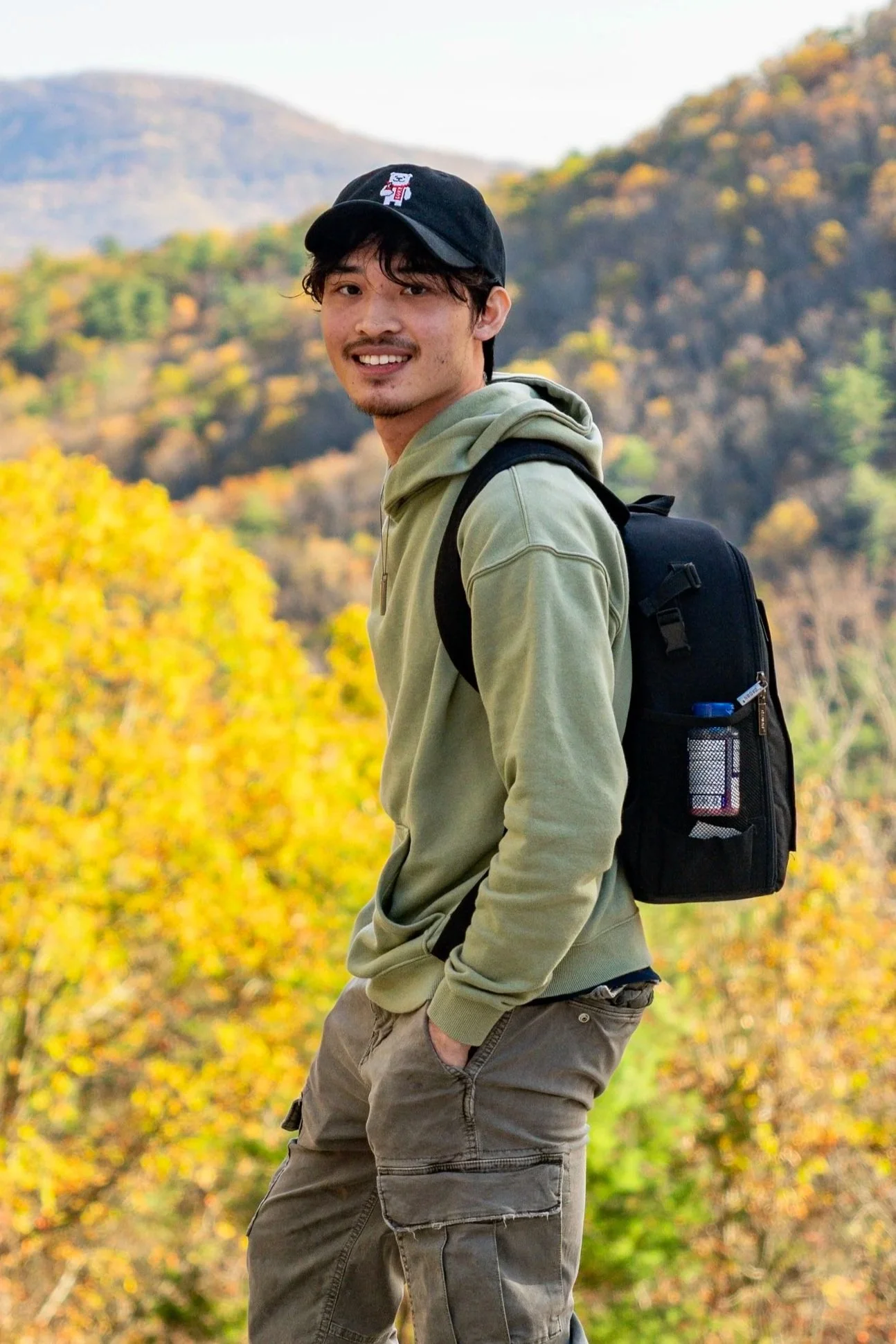 A young man smiling outdoors in front of fall foliage with yellow, orange, and green leaves. He is wearing a black baseball cap with a small graphic, a light green hoodie, and gray cargo pants. He has a black backpack on one shoulder and has one hand in his pocket.