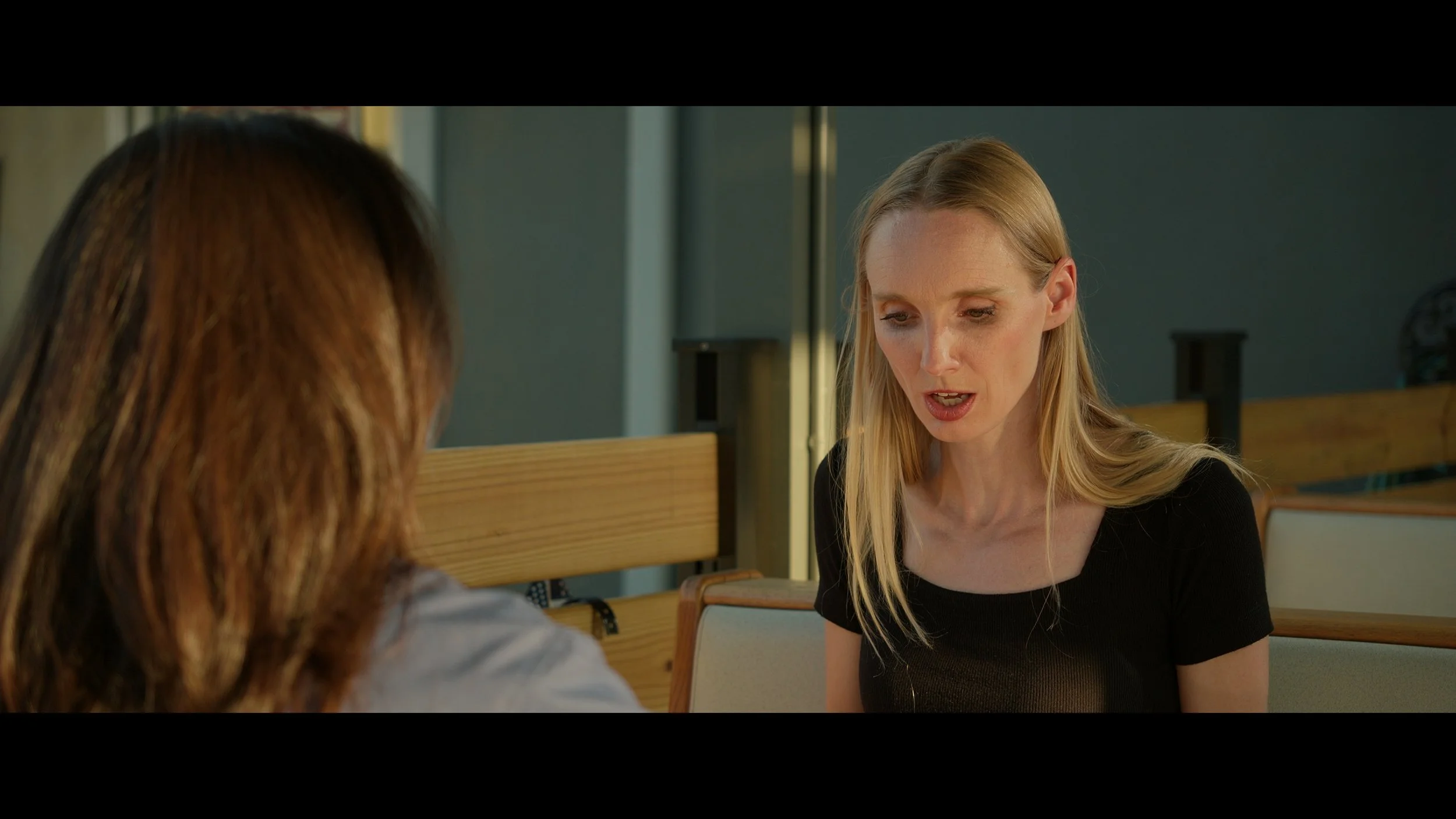 Two women having a conversation in a brightly lit indoor setting, with one woman with long blonde hair and black top speaking to another woman with brown hair, partially visible.