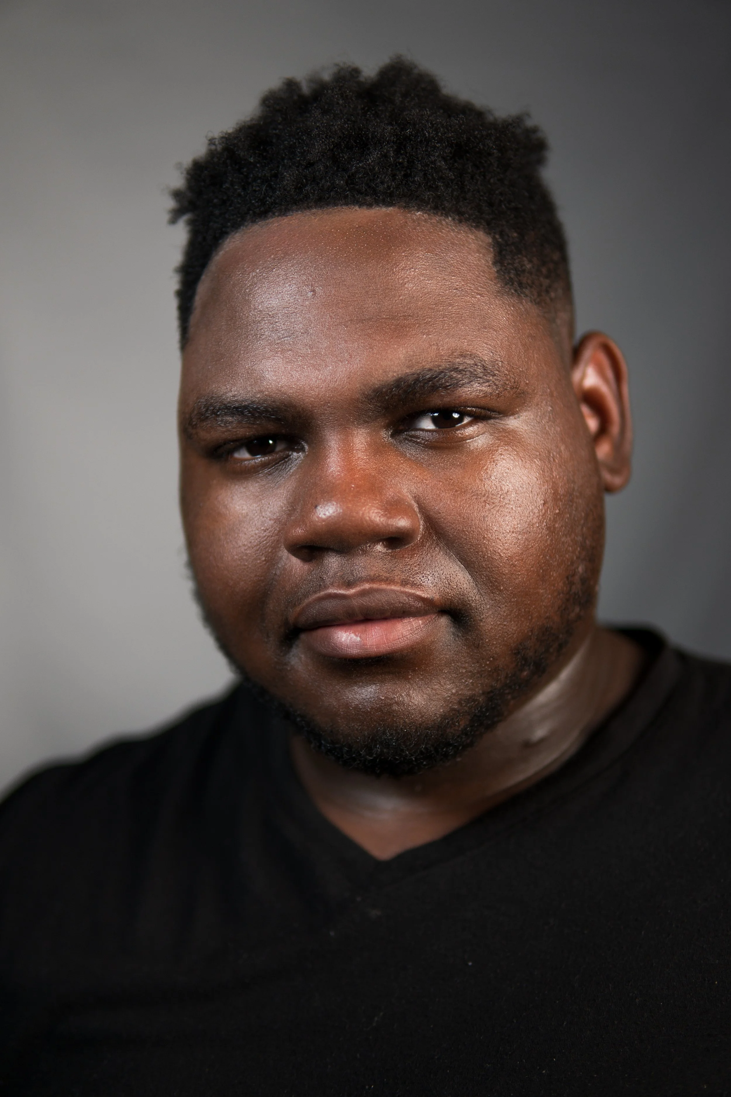 Close-up portrait of a young man with dark skin, short curly hair, and a slight smile. He is wearing a black V-neck shirt, and the background is plain and gray.