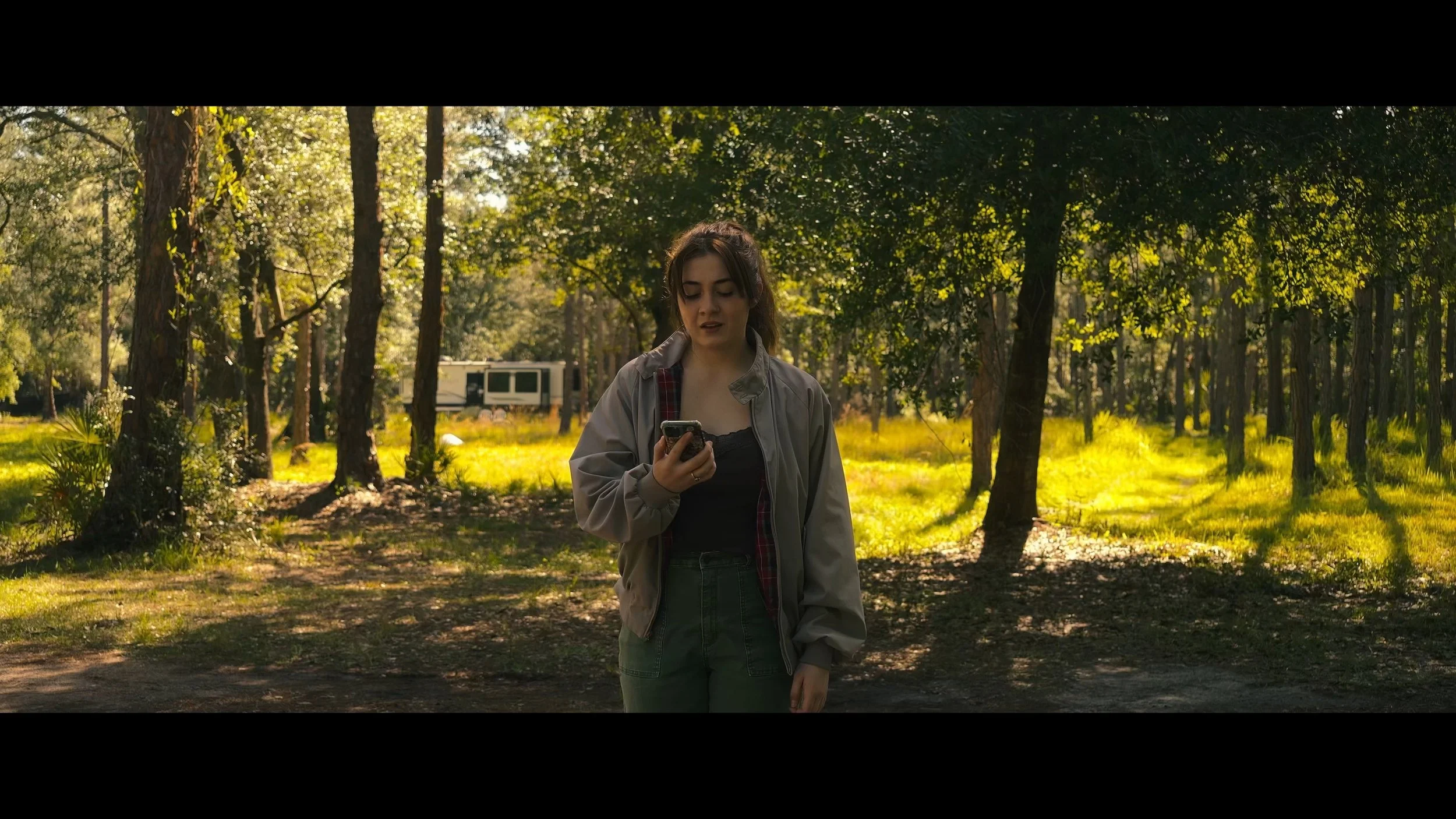 A young woman standing in a sunlit forest, looking at her phone.
