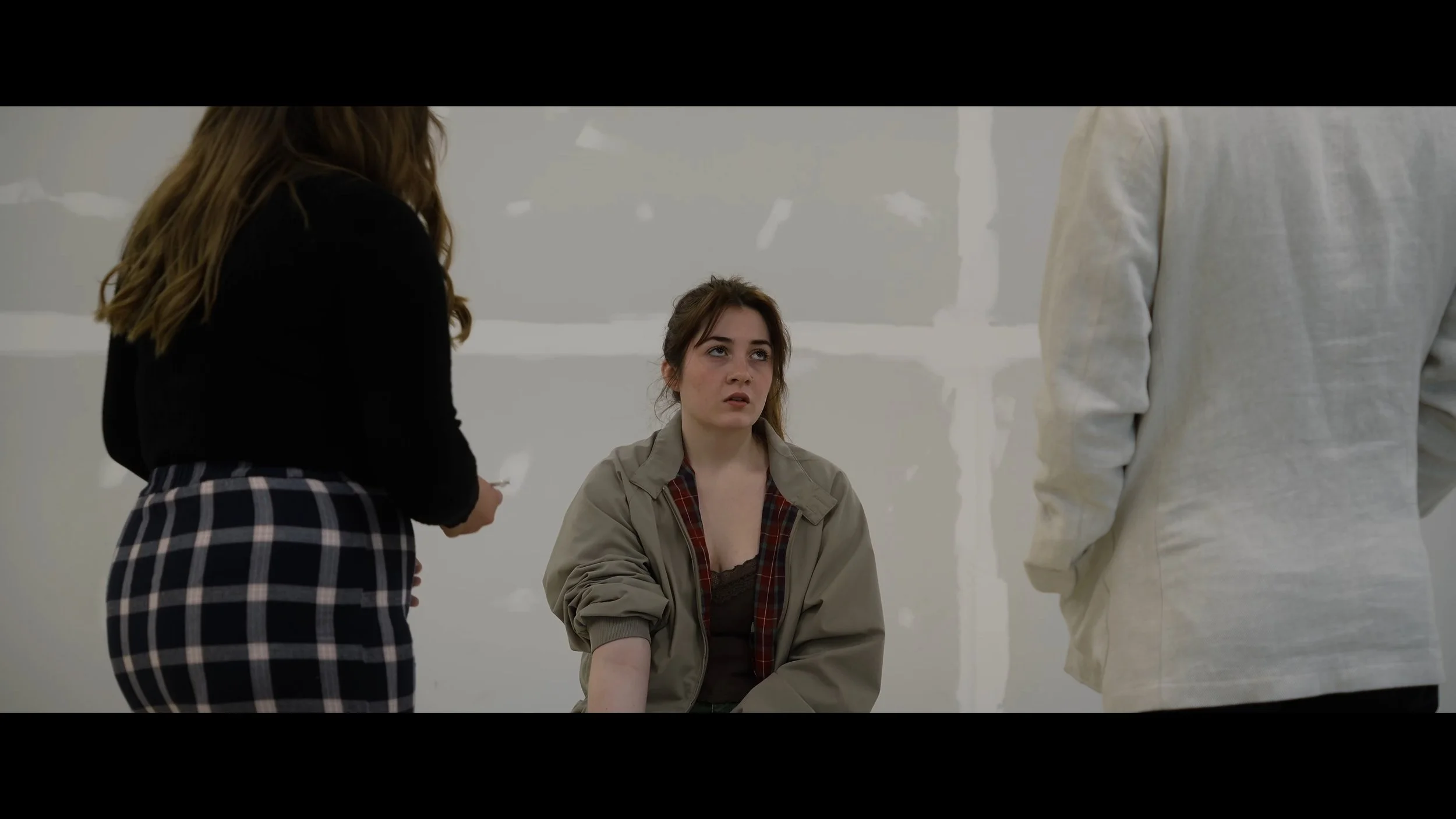 A young woman sitting with a serious expression while three other people stand nearby in a plain, white-walled room.