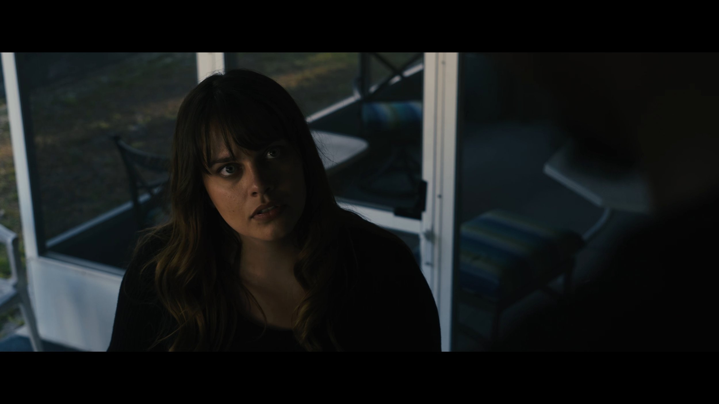 A woman with long brown hair and blue eyes looking intently at something or someone outside the frame. She is indoors, near a glass display case or enclosure with a black frame, and there are chairs and a table inside the enclosure. The background ou