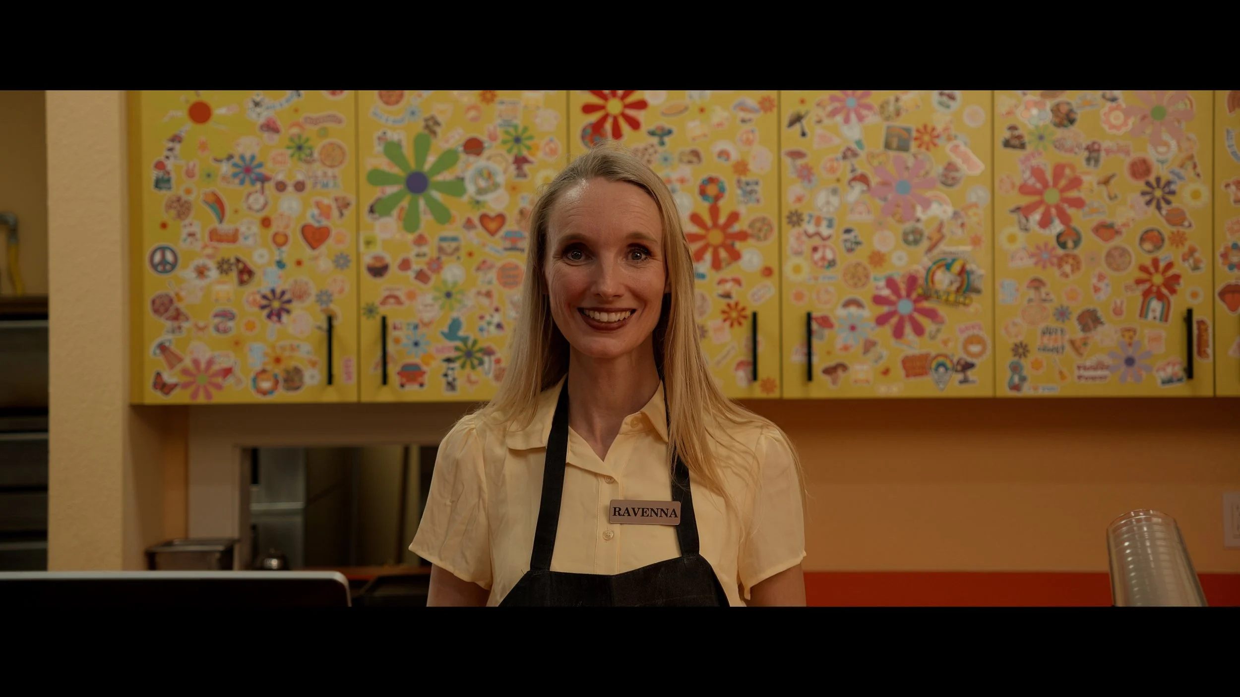 A woman with long blonde hair smiling behind a counter, wearing a yellow shirt and black apron with a name tag that says 'RAVENNA', in front of a colorful, sticker-covered yellow wall.