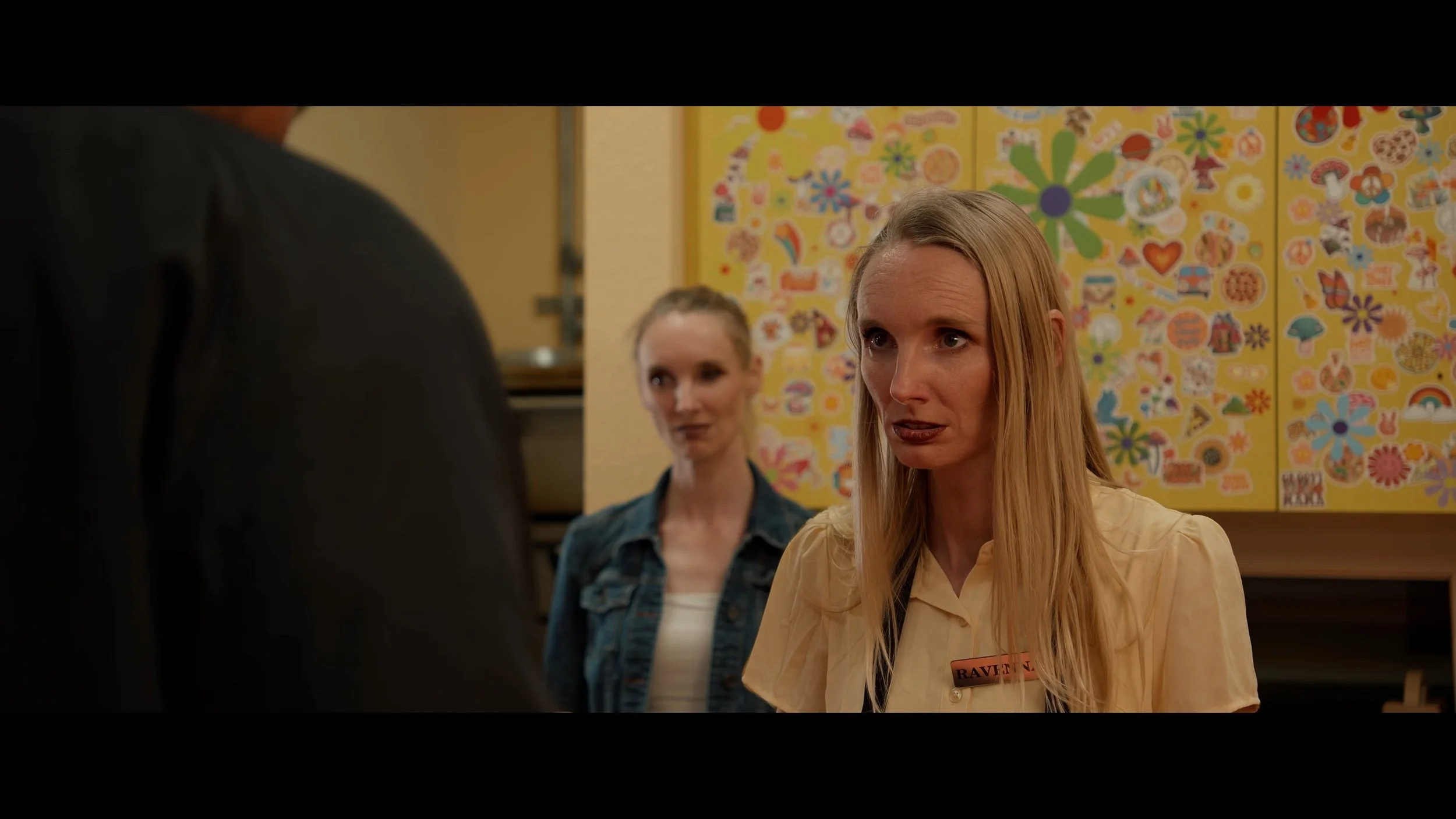 Two women are standing inside a room with a colorful, patterned wall in the background. One woman has red hair, wearing a beige shirt with a name tag, and is speaking towards a person in a dark shirt. The other woman, with lighter hair tied back, is 