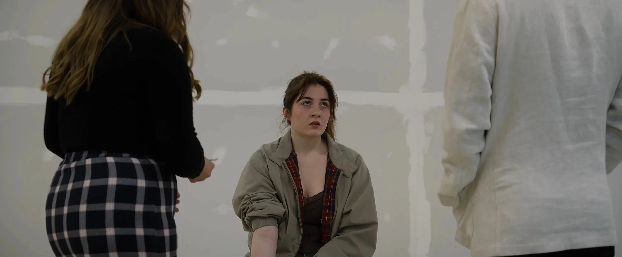 A young woman sitting on the floor with an annoyed or tired expression, surrounded by two other people standing around her. One person is a woman with long, wavy hair wearing a black top and plaid skirt, and the other person is partially visible, wearing a light-colored jacket.